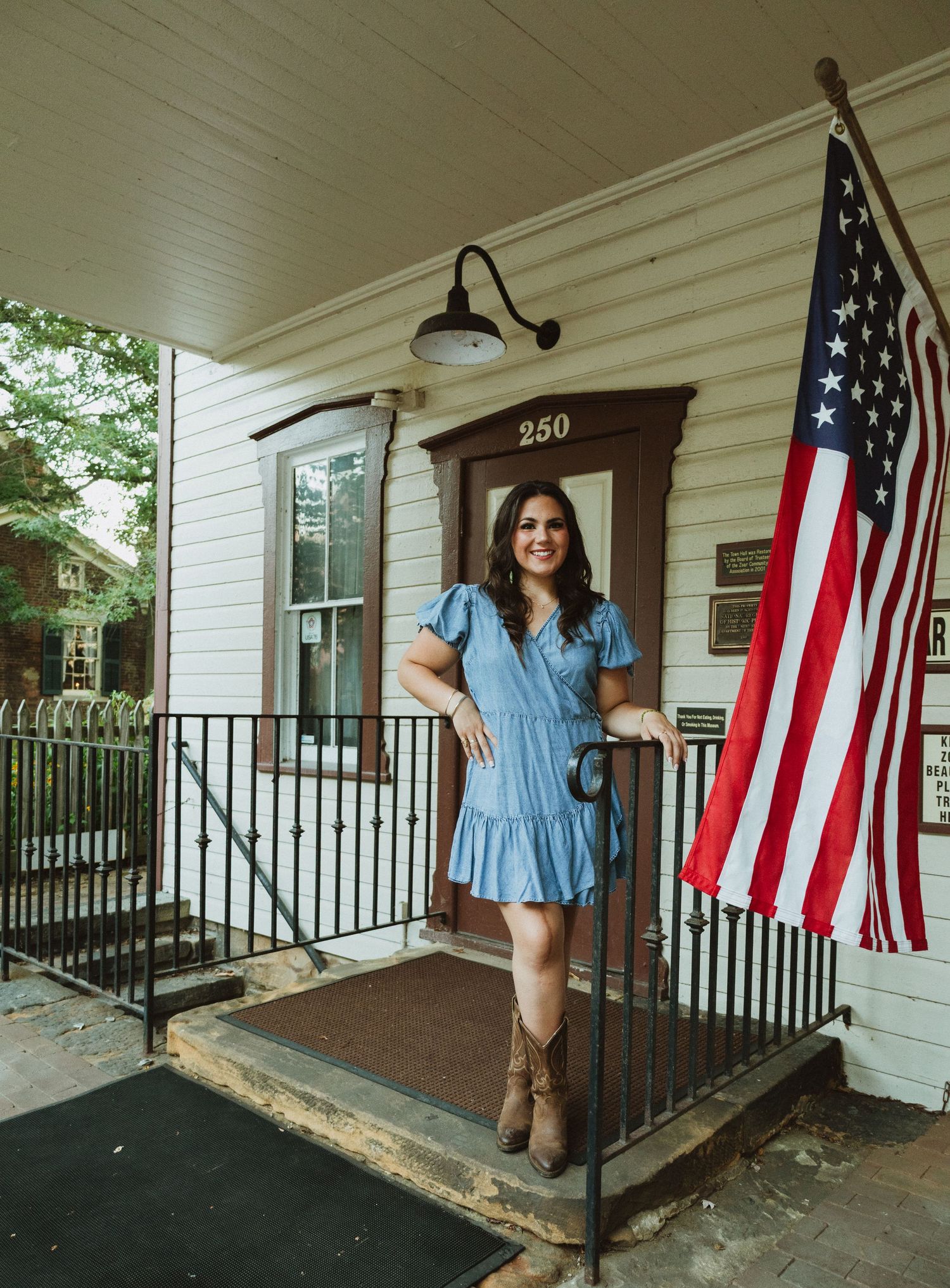 A classic white porch with American flag and black railings features someone in a blue dress posing by the doorway.