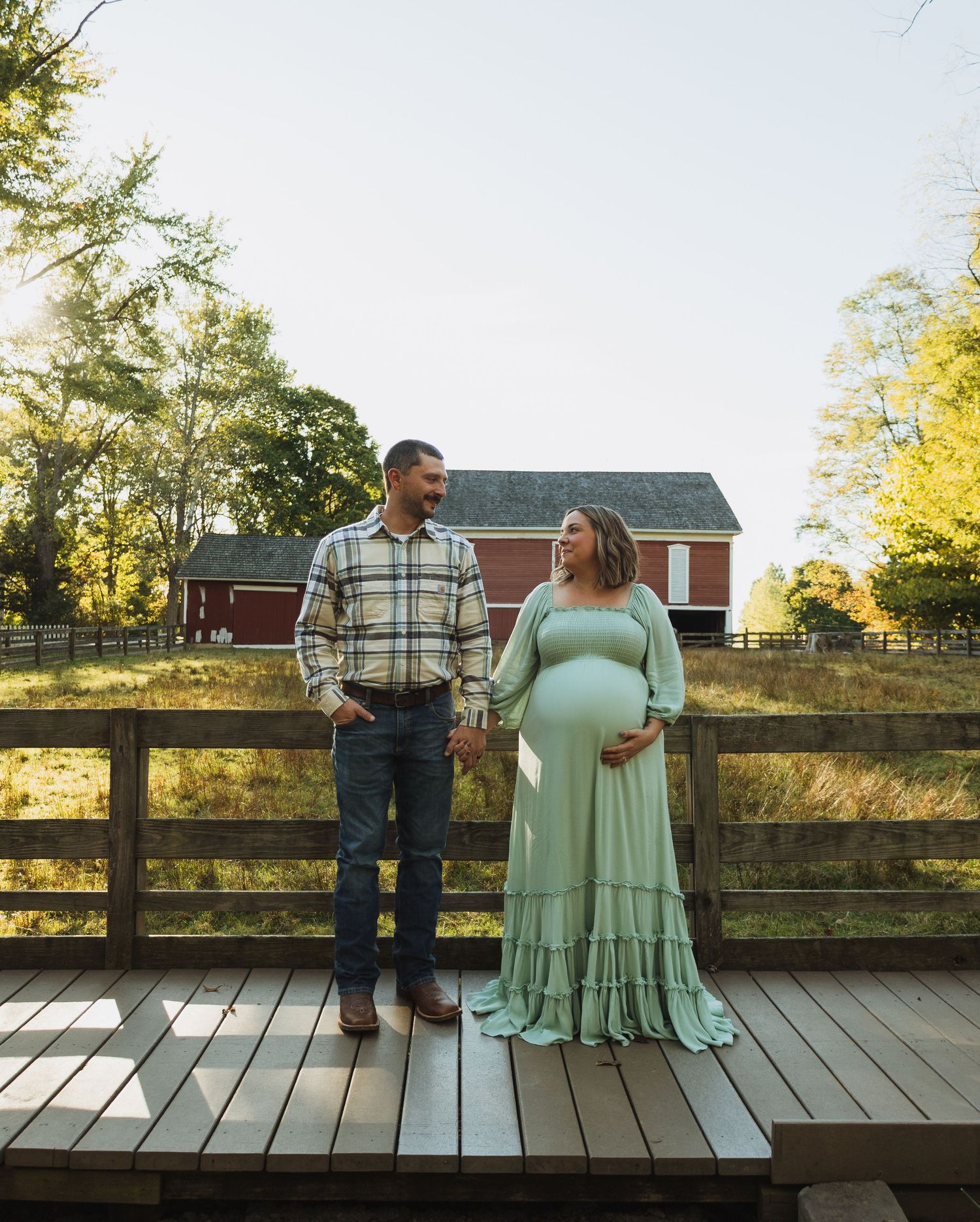 A couple stands on a wooden deck with a red barn in the background during a maternity photoshoot in golden sunlight.