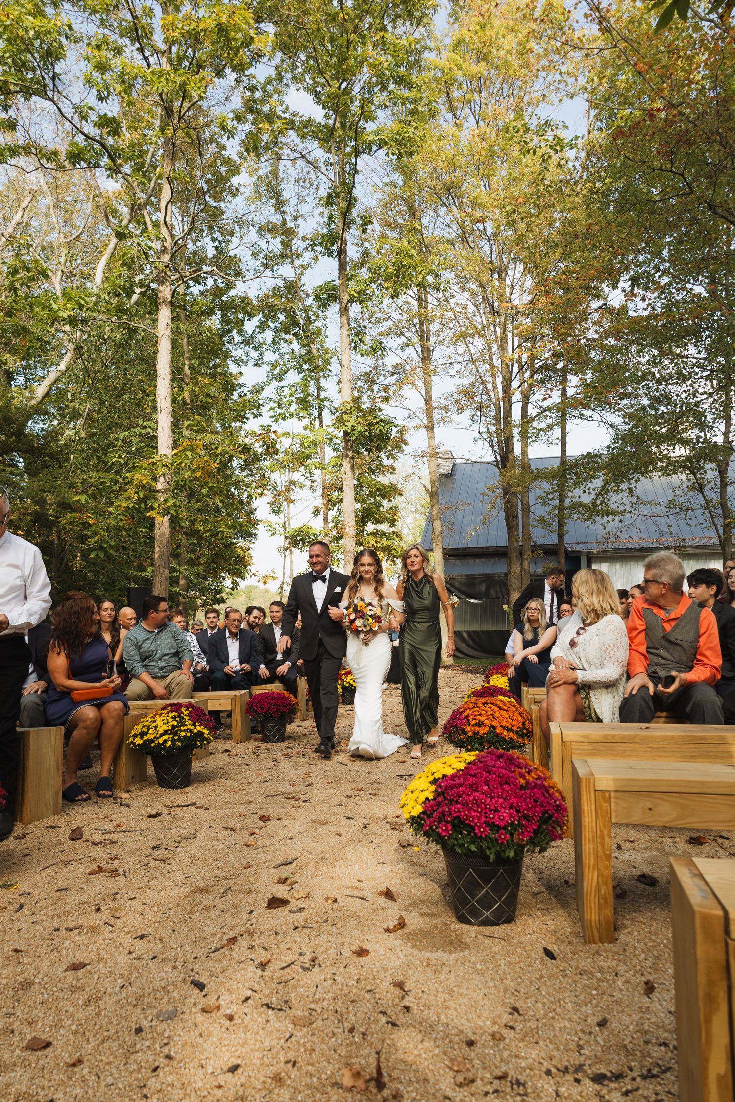 Outdoor wedding ceremony setup with wooden benches and colorful mums surrounded by autumn trees.