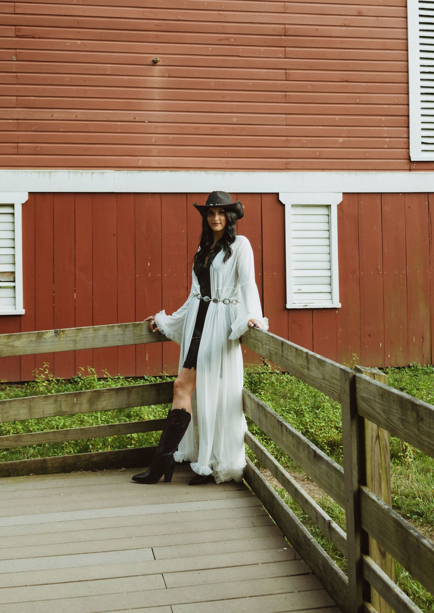 Person in traditional dress and black bonnet stands on a wooden boardwalk beside a red barn.