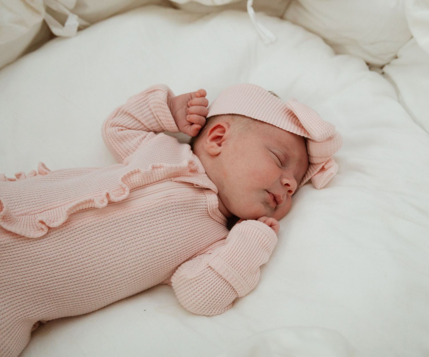 A sleeping newborn baby dressed in a soft pink ribbed outfit rests peacefully on white bedding.
