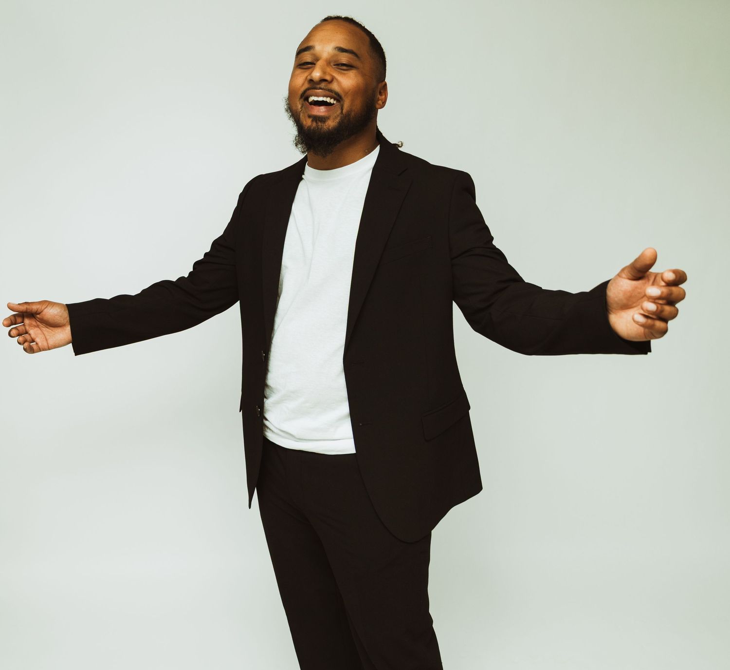 A person in a black suit and white t-shirt poses with arms spread wide against a light background.