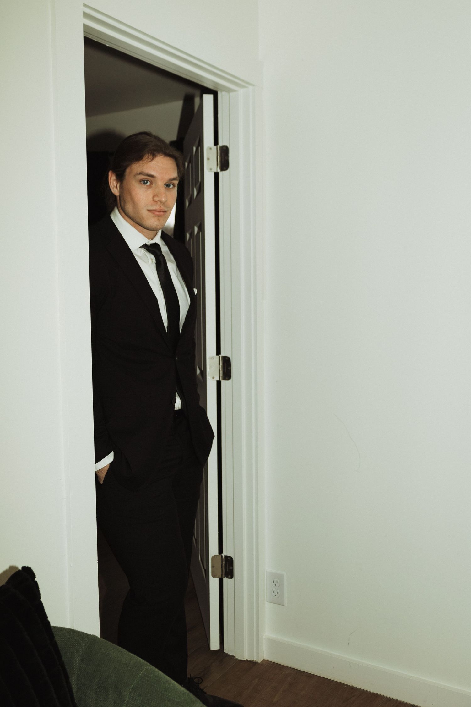 Person in black suit and tie stands in doorway with hand in pocket.