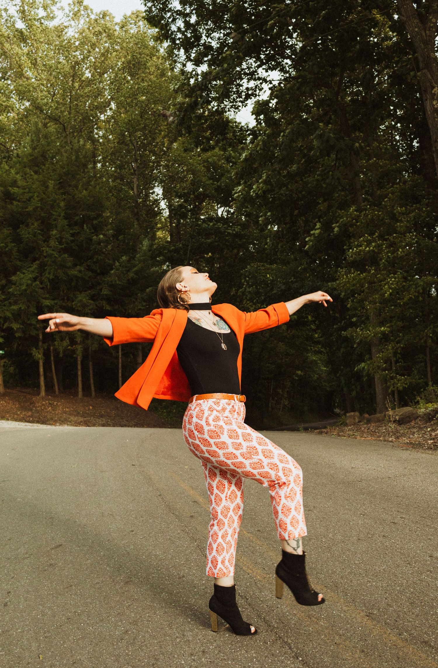 Dynamic fashion pose on road showing orange blazer and patterned pants with arms outstretched.