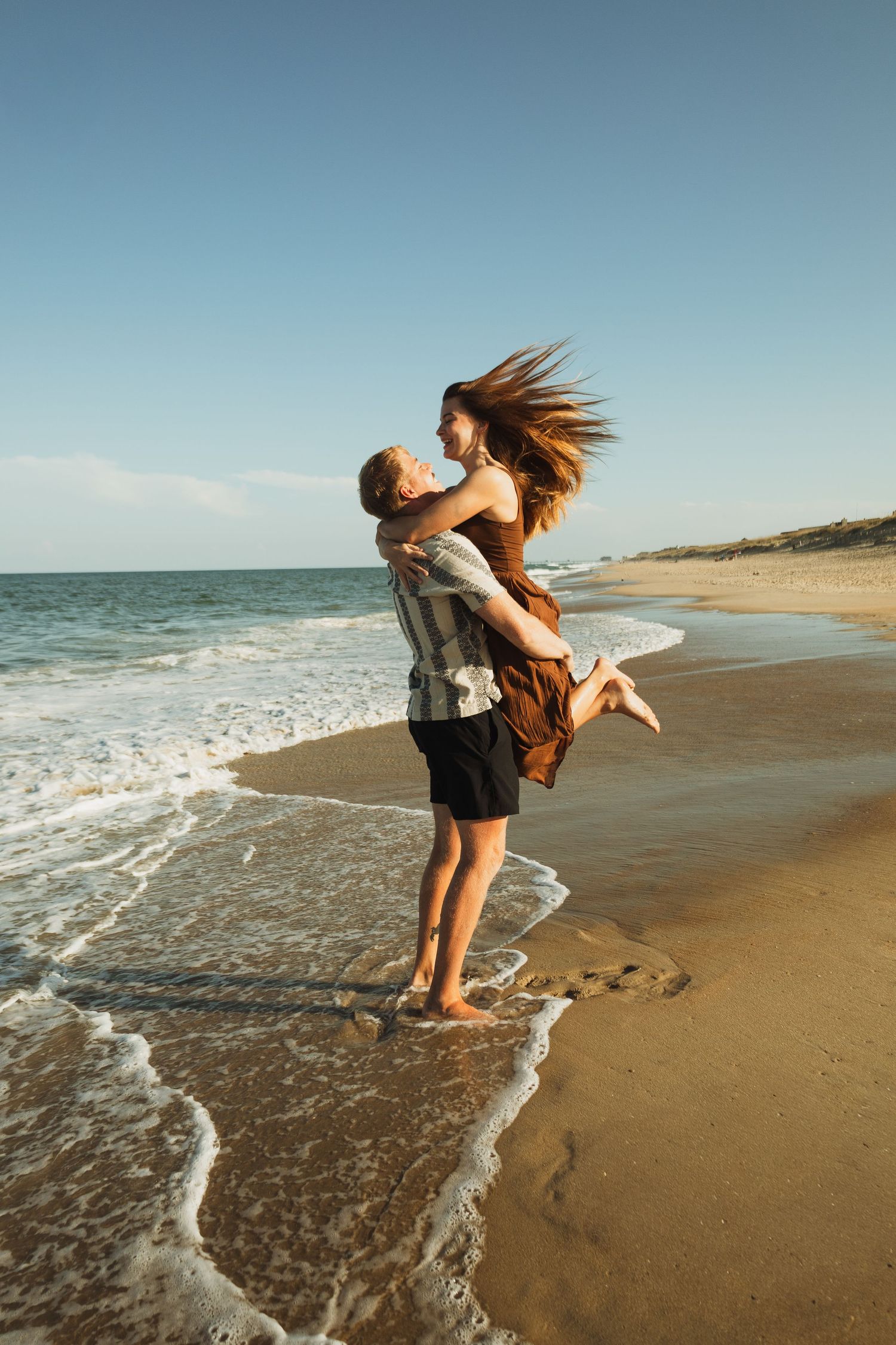 Playful couple embracing at the beach during sunset with waves crashing at their feet.