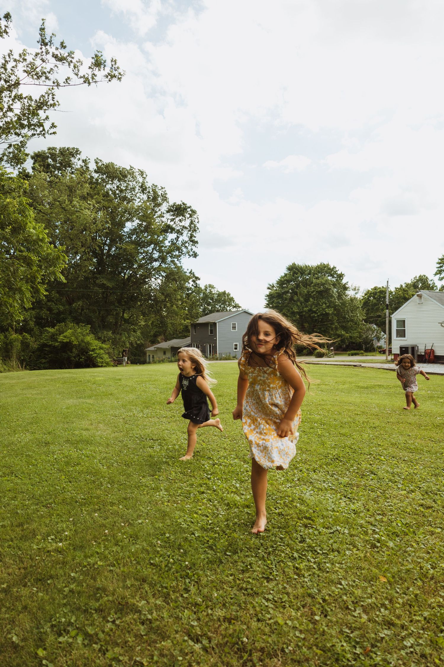 Three young girls running and playing on a green lawn surrounded by trees and houses on a sunny day.