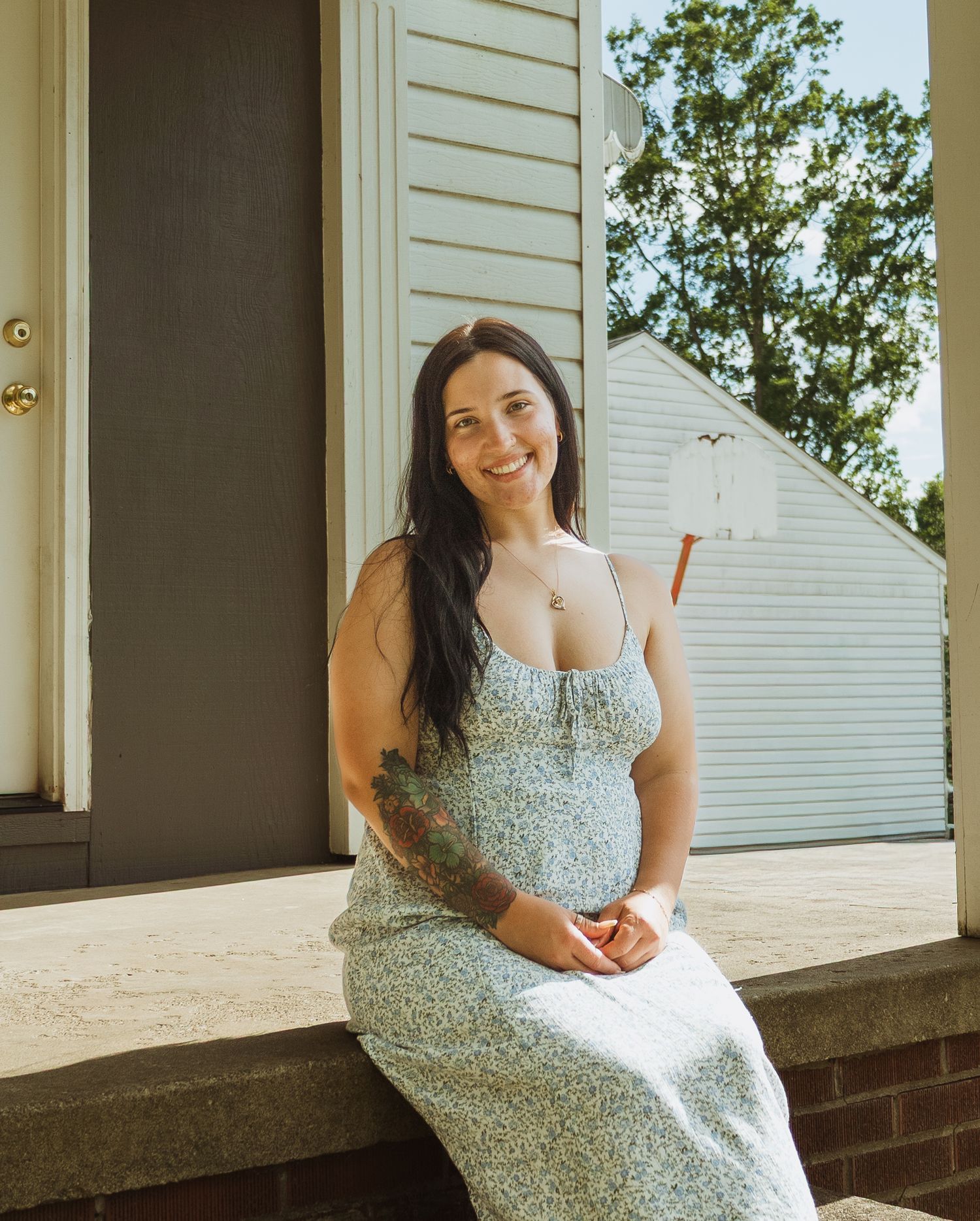 Smiling pregnant woman in floral dress sitting on porch steps with tattoo, long dark hair, sunny day.