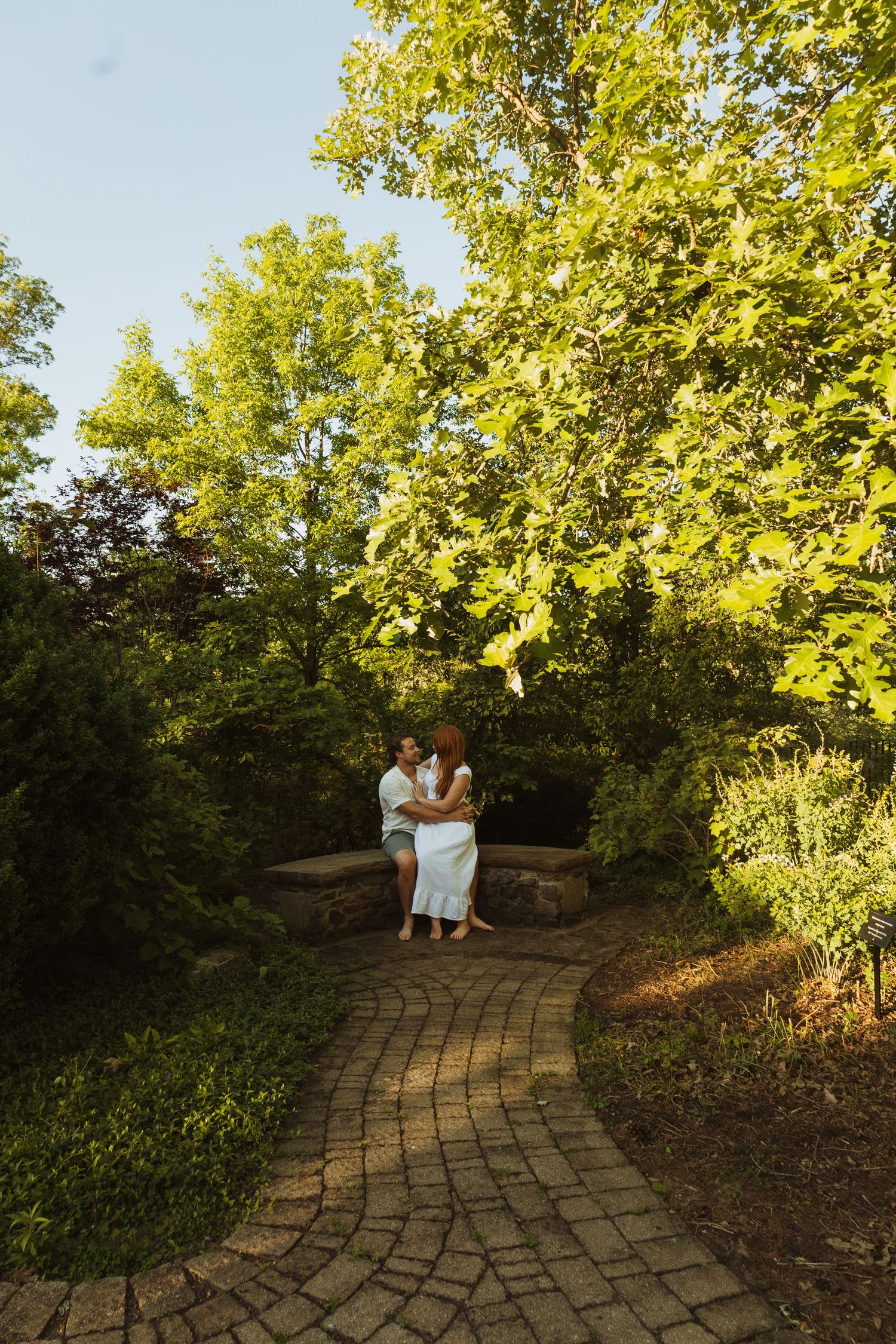 Couple embracing on a brick garden path surrounded by lush green trees in a romantic outdoor setting.