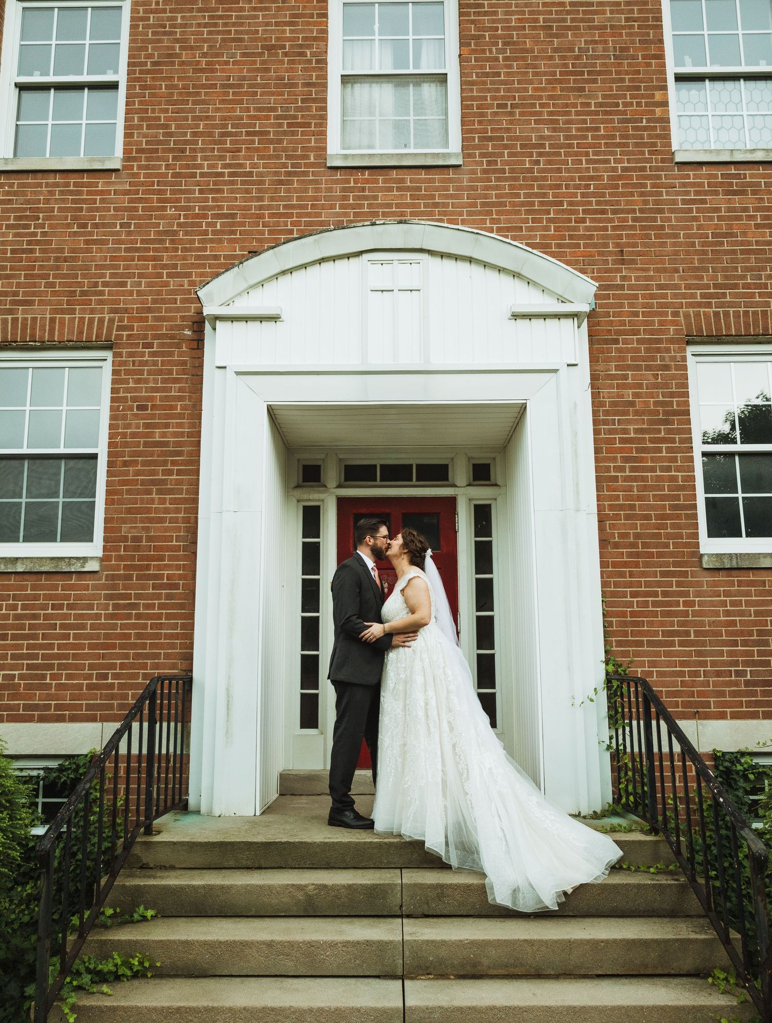 A wedding couple poses on the steps of a classic red brick building with white trim and columns.
