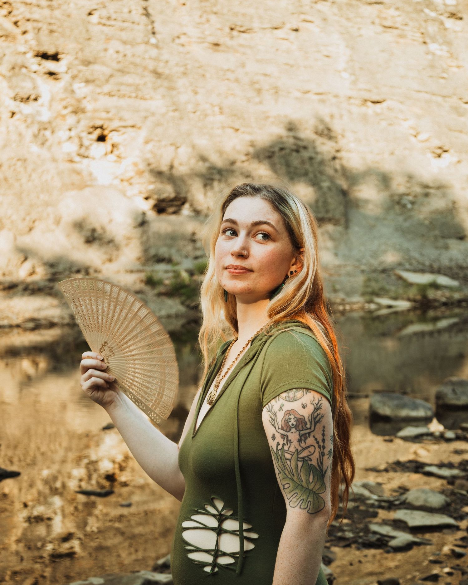Woman with tattoos poses outdoors in green top while holding a decorative hand fan against rocky cliff backdrop.