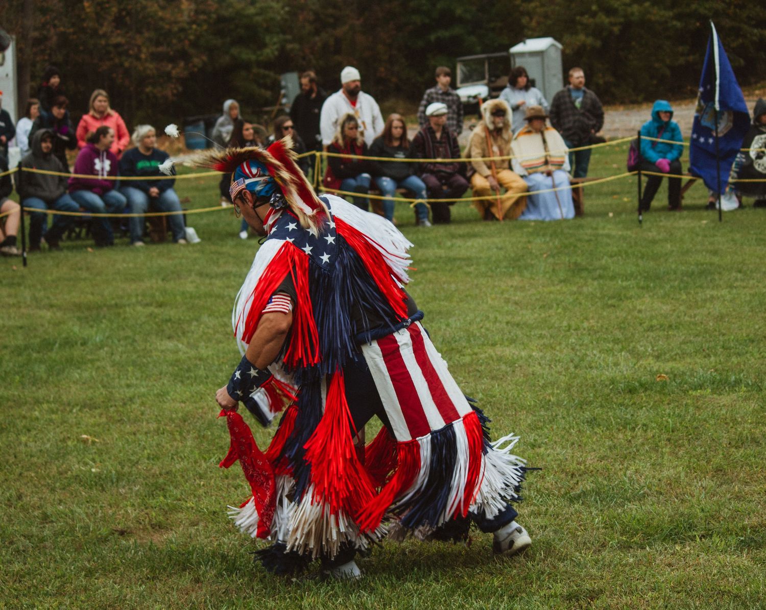 A traditional Native American dancer in regalia featuring American flag patterns performs at an outdoor powwow.