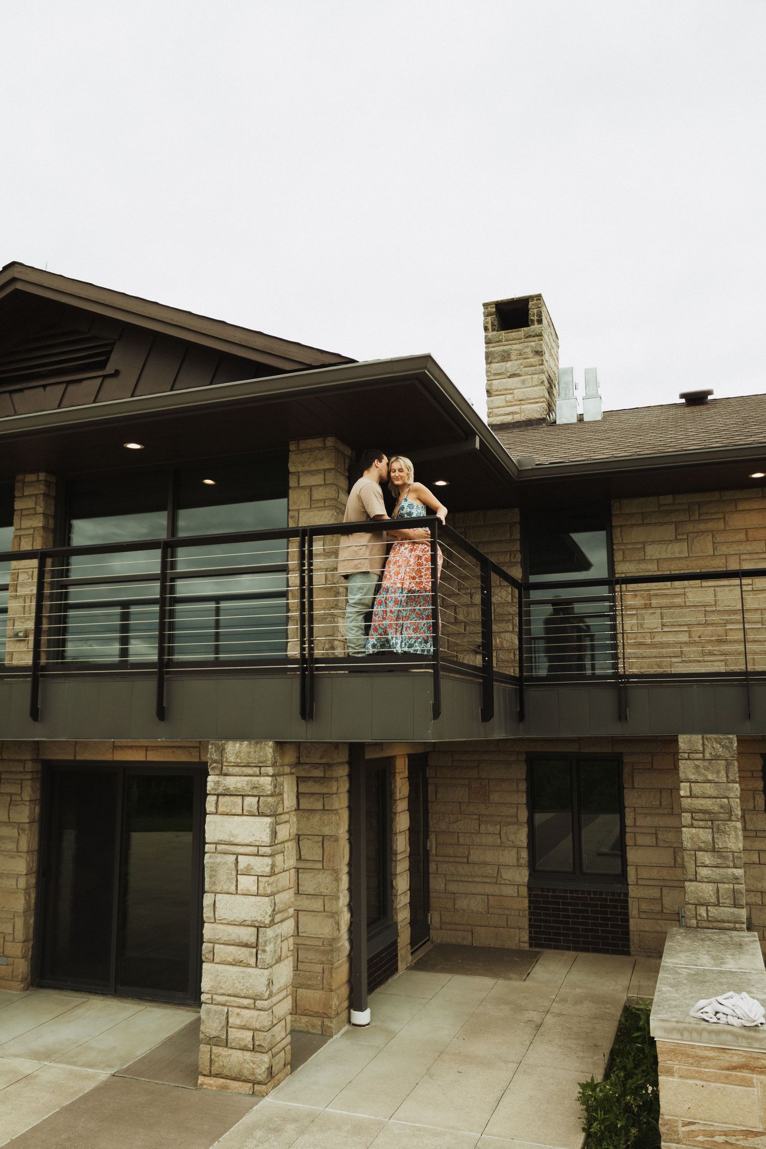 A couple shares an intimate moment on a modern stone house balcony with contemporary architecture.