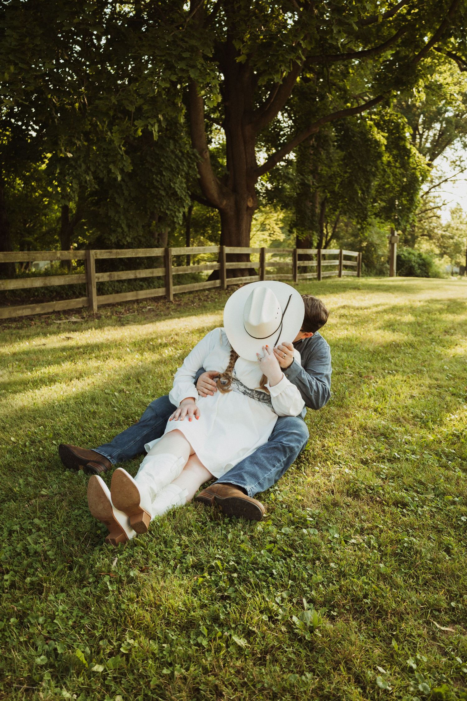 Two people lie together on a grassy hillside during golden hour at a park with wooden fence in background.