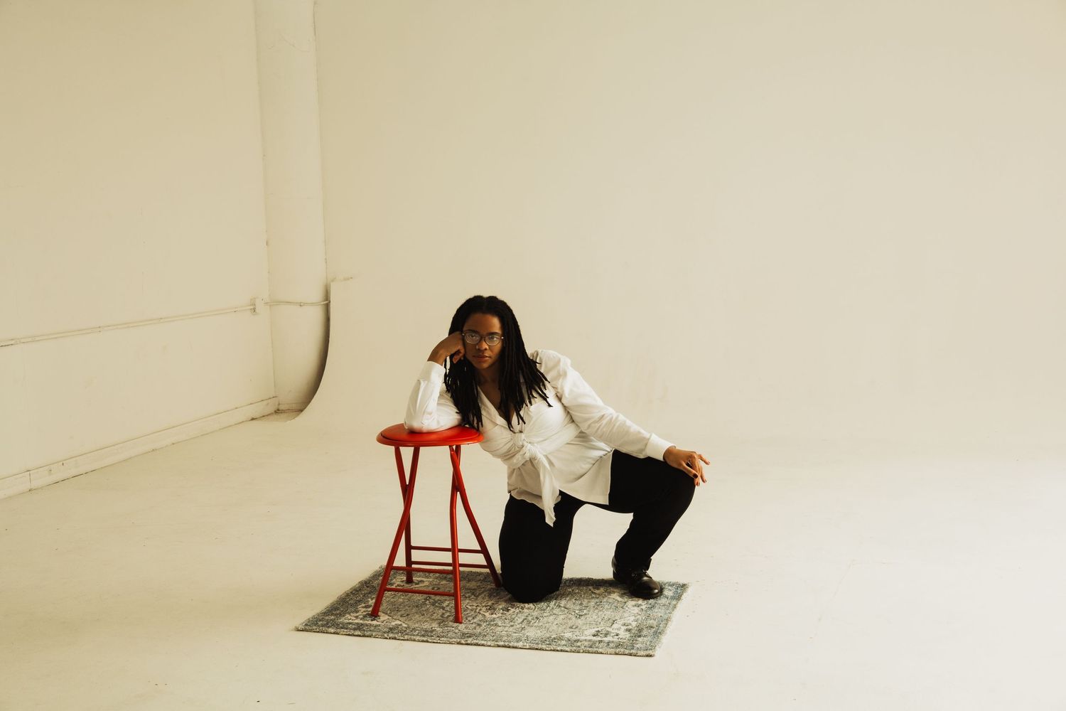 Figure in white jacket sits on red stool against white studio backdrop with decorative carpet underneath.