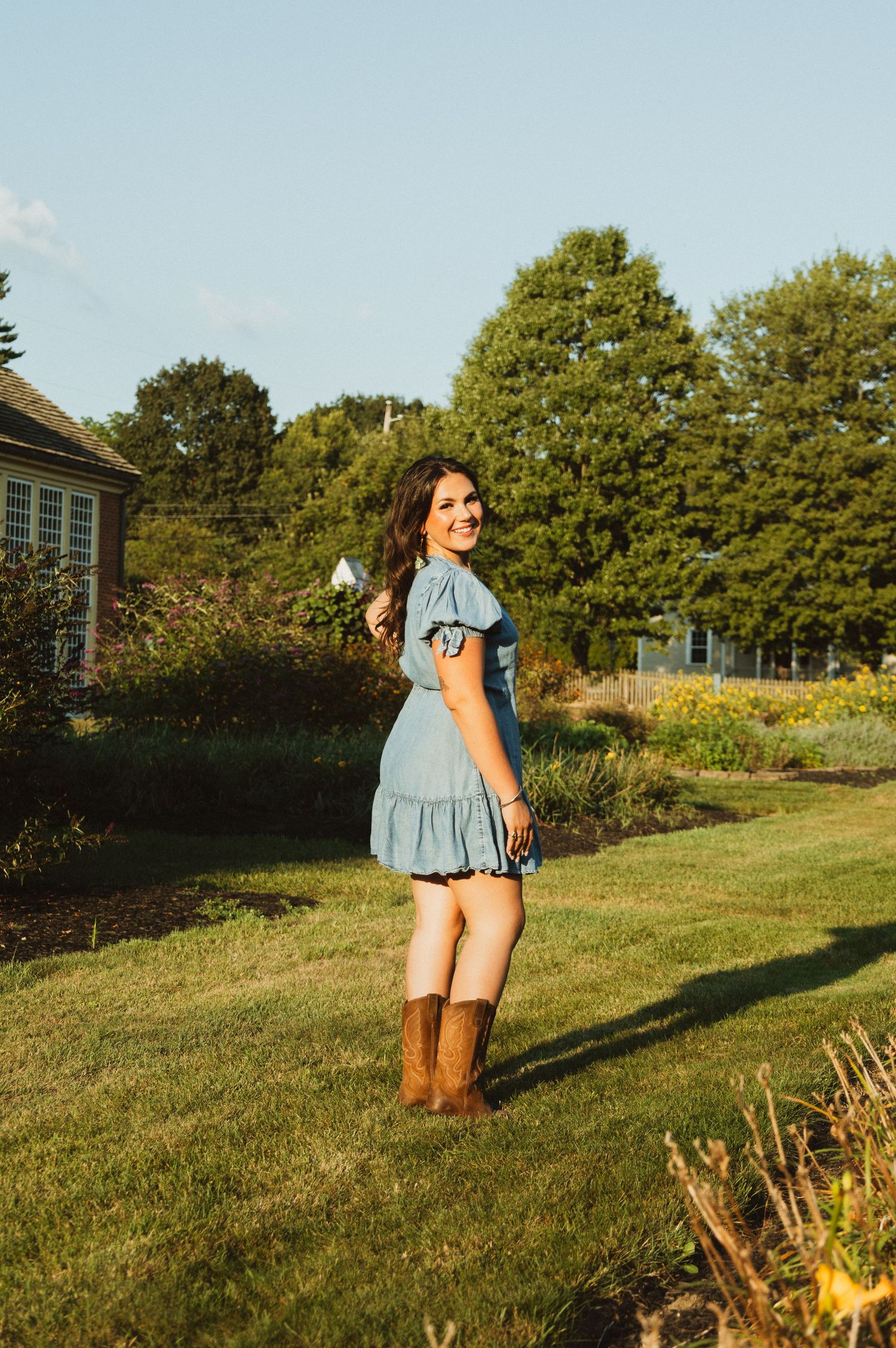 A person wearing a blue dress and tan boots poses casually on a grassy hillside on a sunny day.