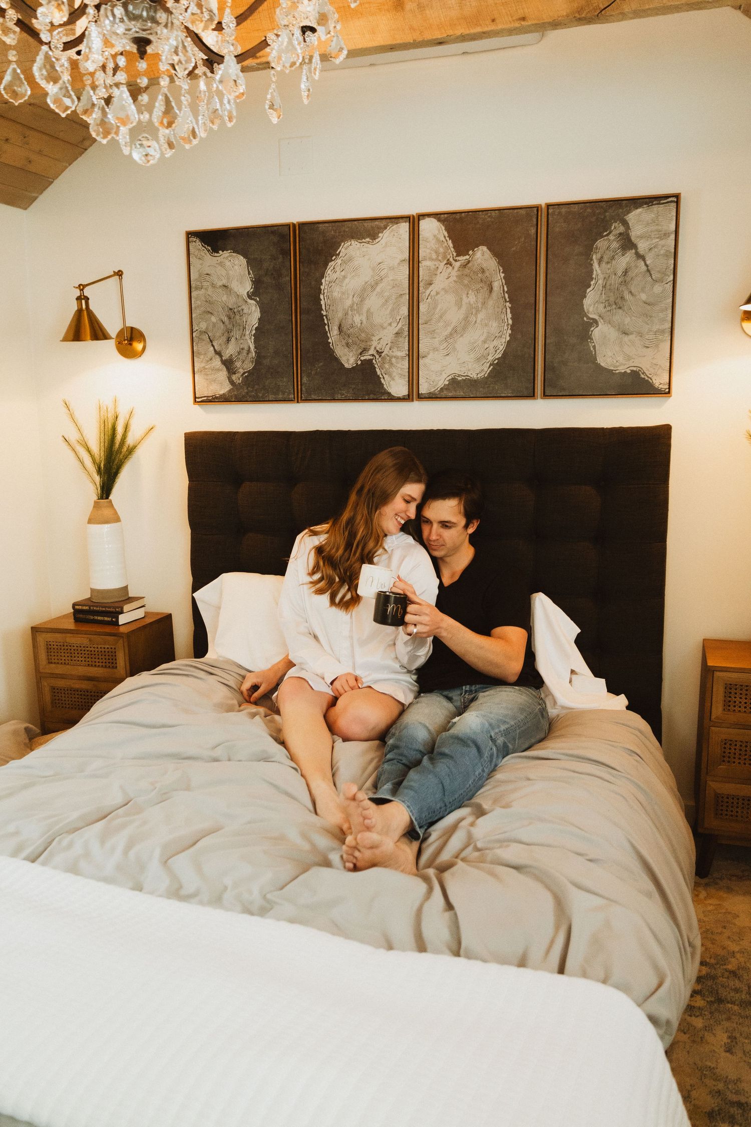 Cozy bedroom scene with abstract wall art, crystal chandelier, and couple sharing intimate moment on grey bedding.
