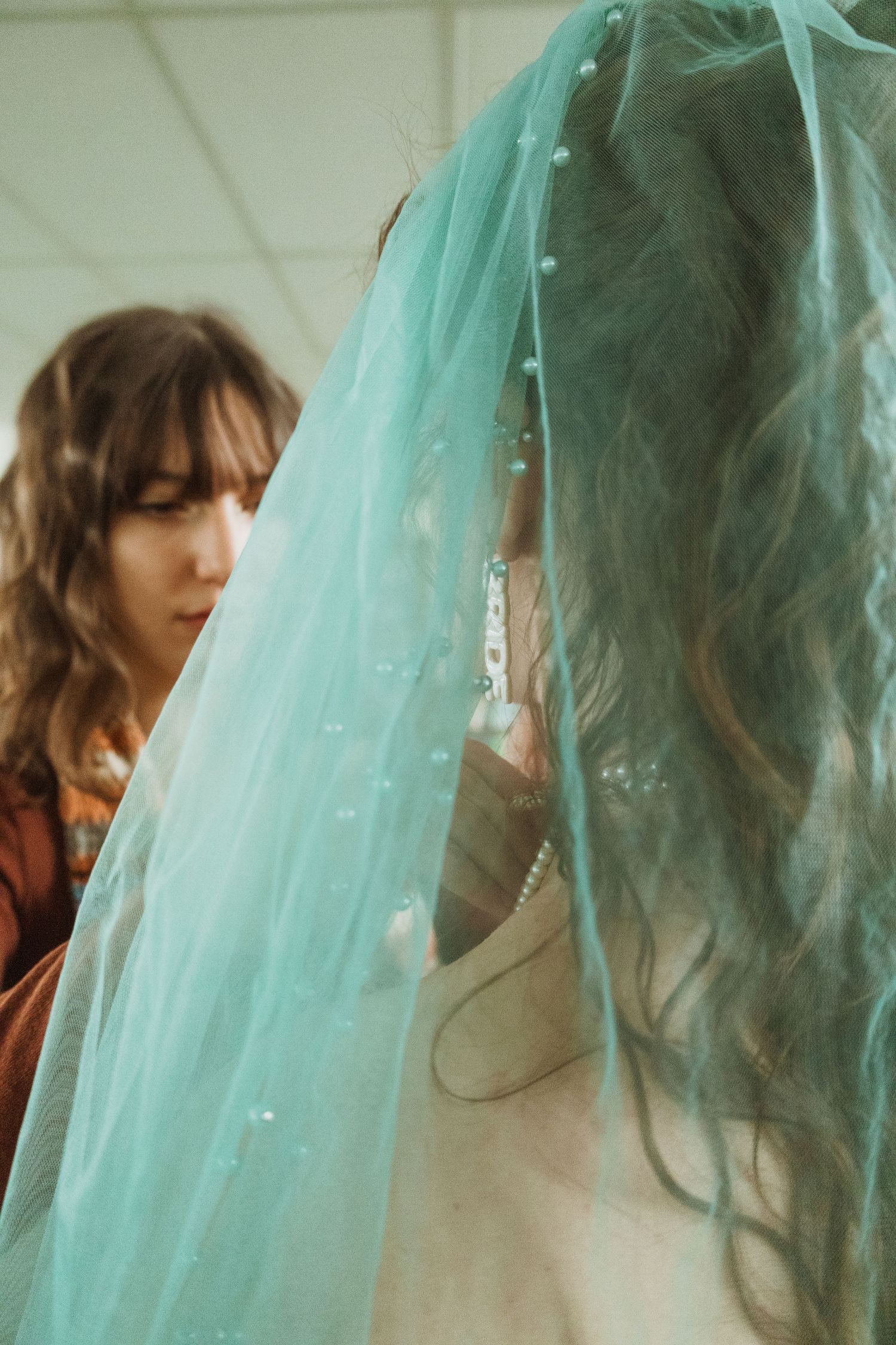 A bridesmaid adjusts a teal tulle veil adorned with pearls on a bride with curly hair.