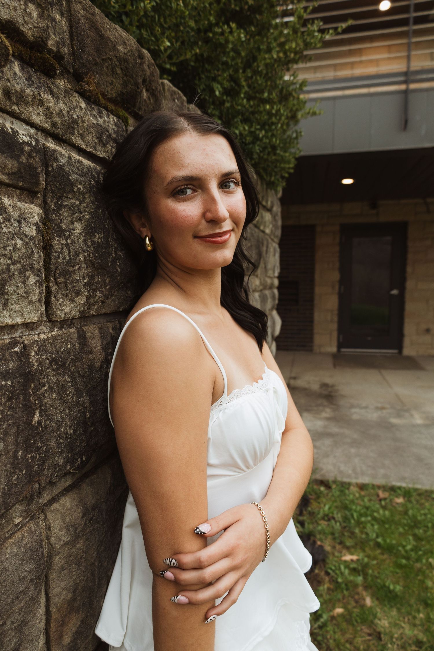 Young woman in white dress leaning against stone wall, smiling outdoors near building.