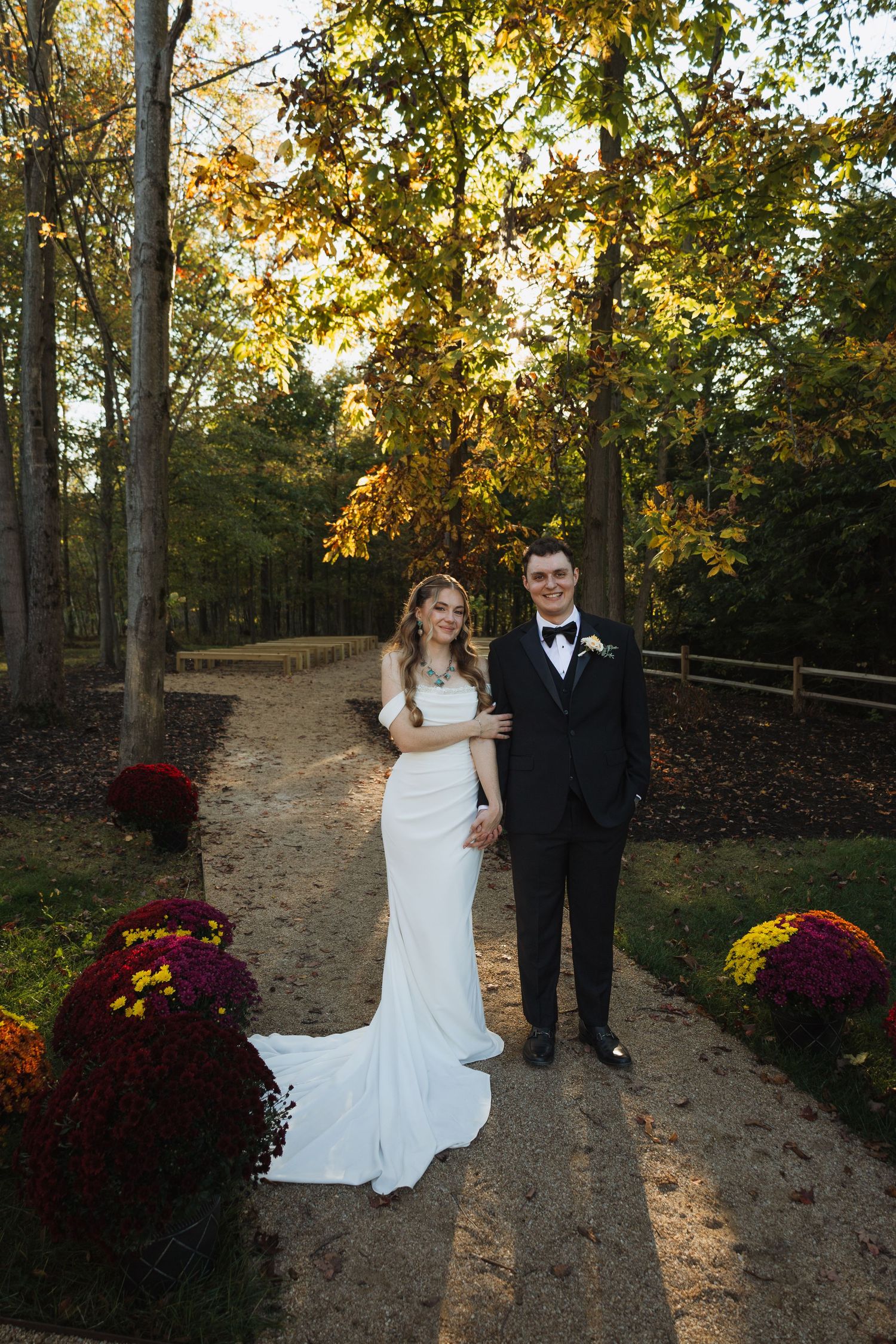 A couple poses in formal attire on a wooded path decorated with burgundy mums during an autumn wedding.