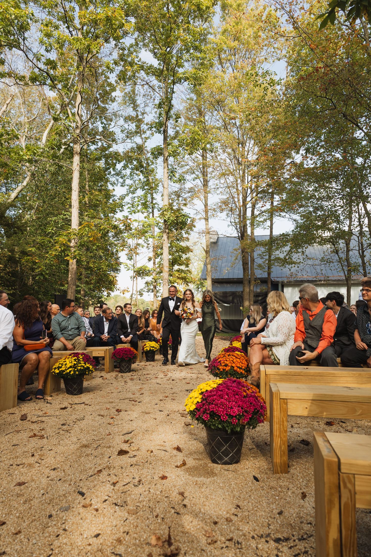 Wedding guests seated on wooden benches surrounded by autumn trees and colorful mums at an outdoor ceremony.