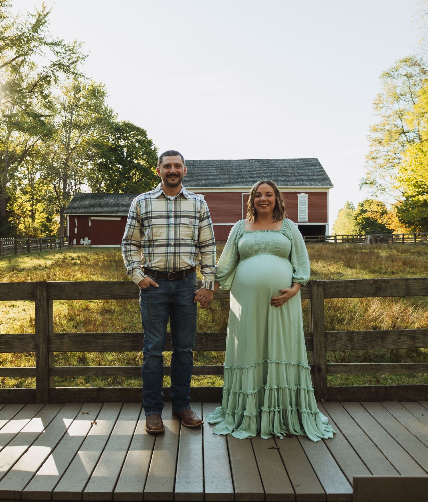 A couple stands together on a wooden deck with a rustic fence and autumn foliage in the background.