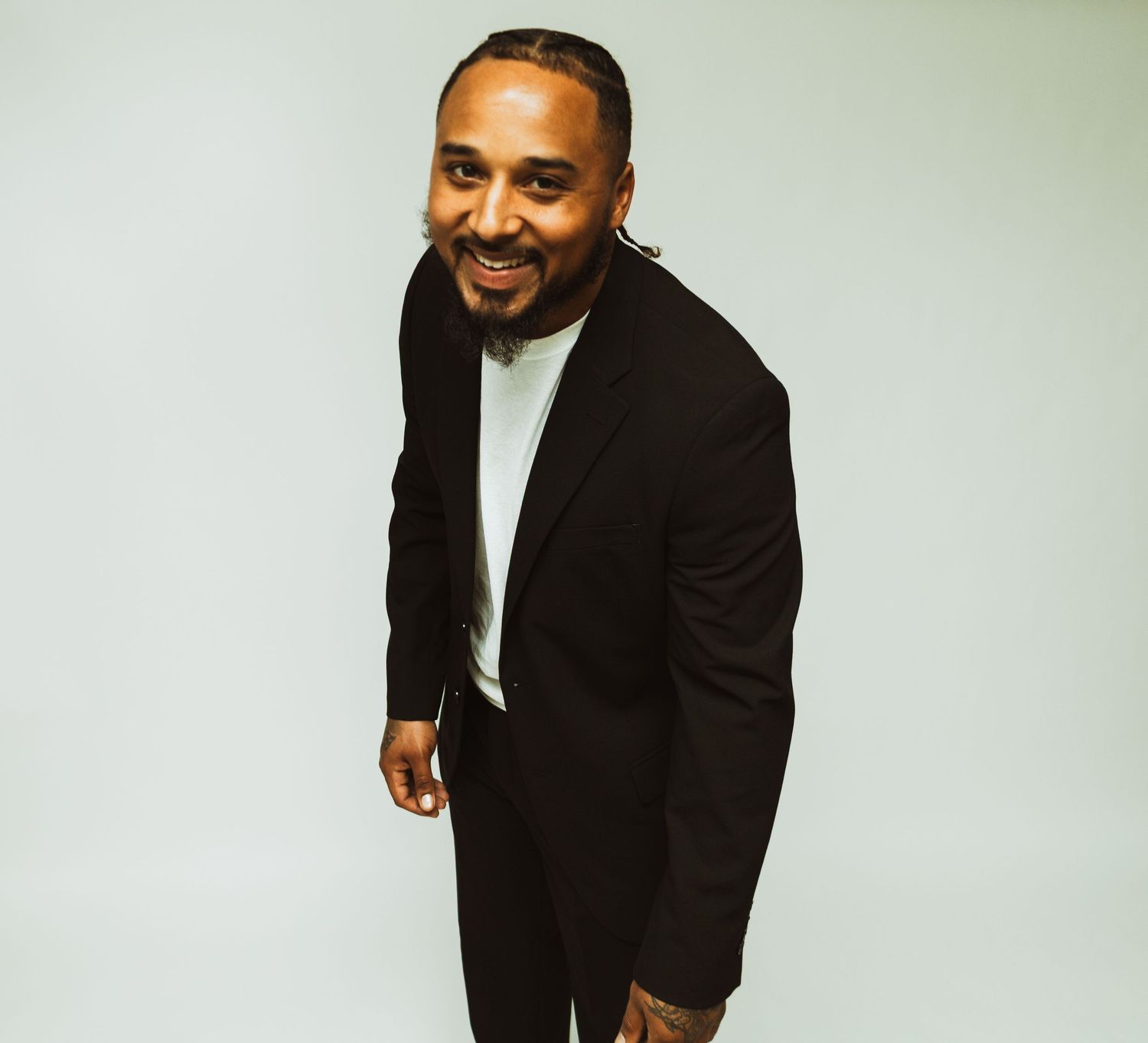 A person in a black blazer and white shirt smiles warmly while leaning forward against a light background.