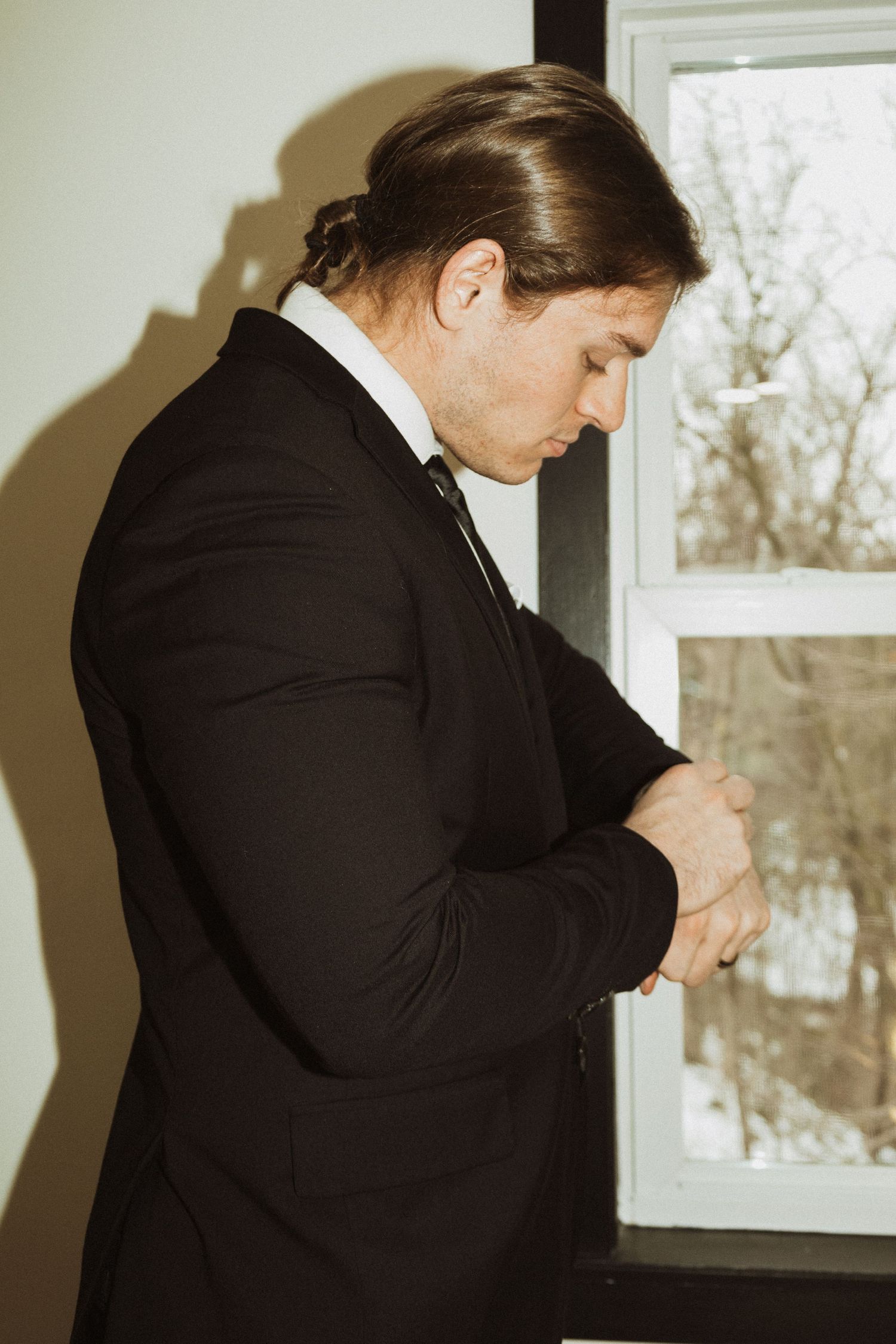 Person in black formal suit adjusts tie while standing near window.