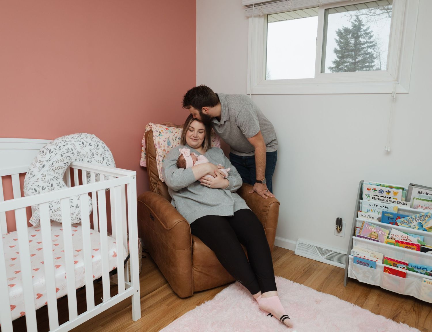 Sweet nursery scene with white crib, brown rocking chair and book display shelf in corner of pink-walled room.