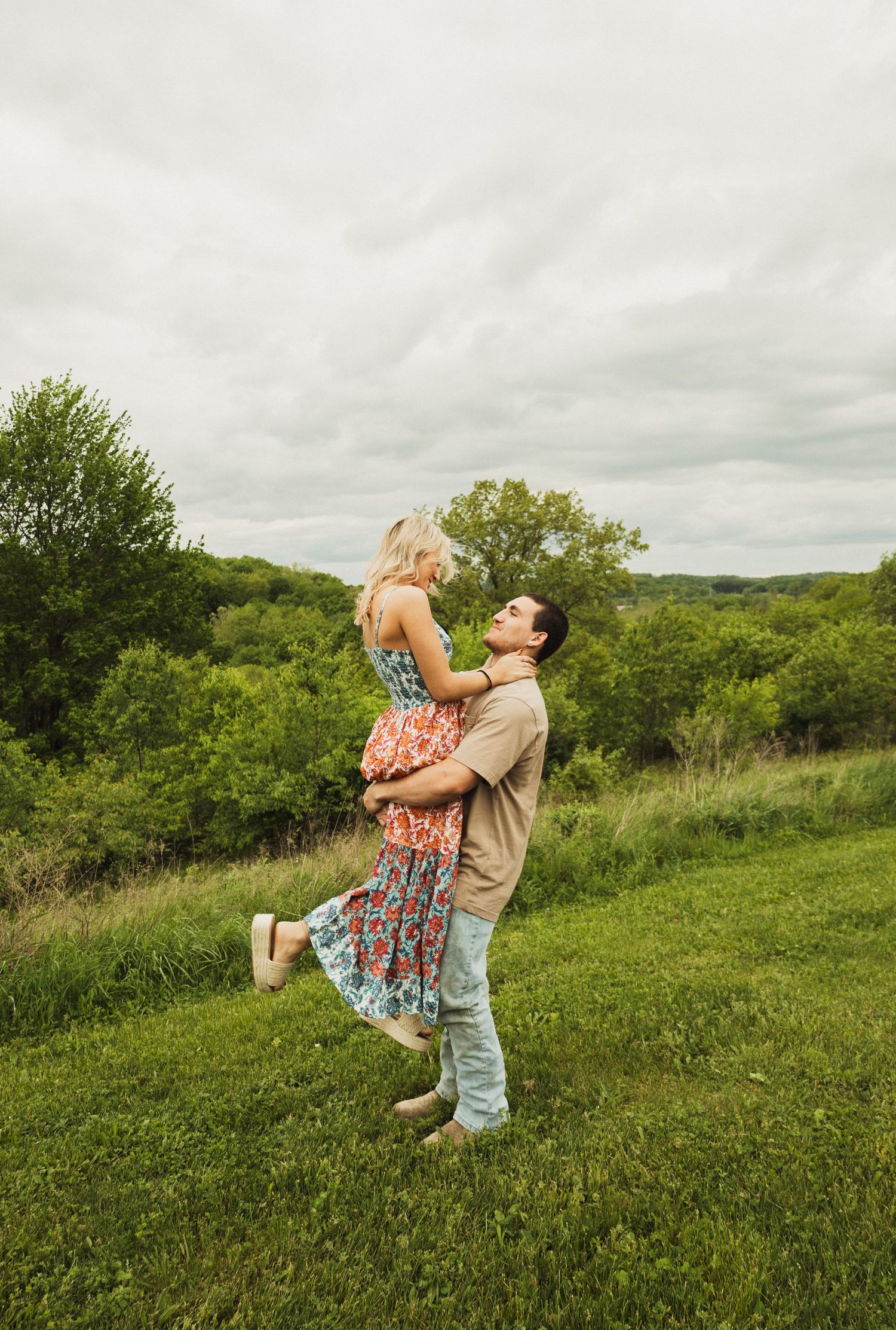 A couple embraces in a romantic lift pose against a scenic green hillside backdrop on an overcast day.