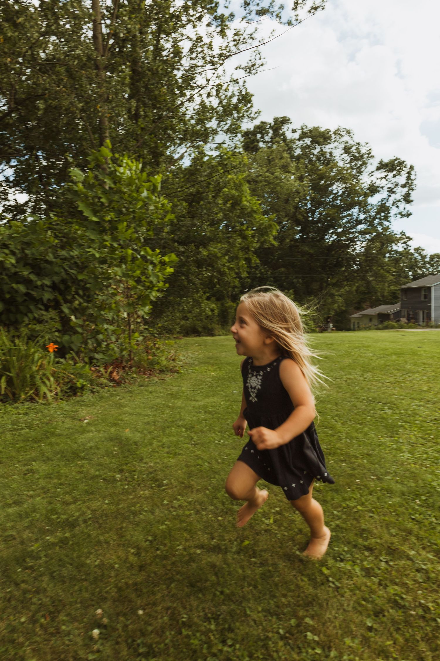 A barefoot young girl in a black dress runs joyfully across a lush green lawn with trees in the background.