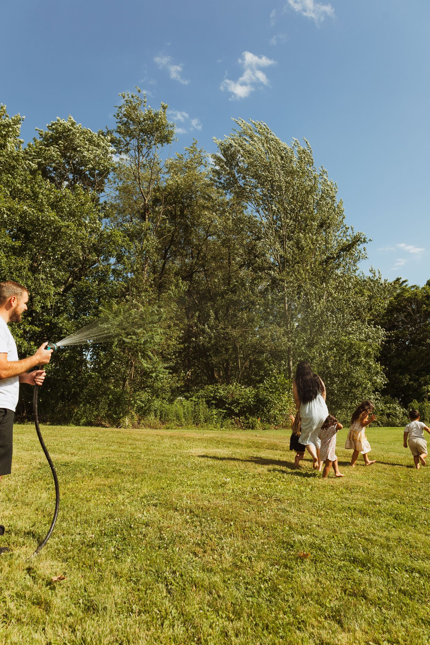 Family playing with garden hose on sunny day, father spraying water at mother and children running on green lawn.
