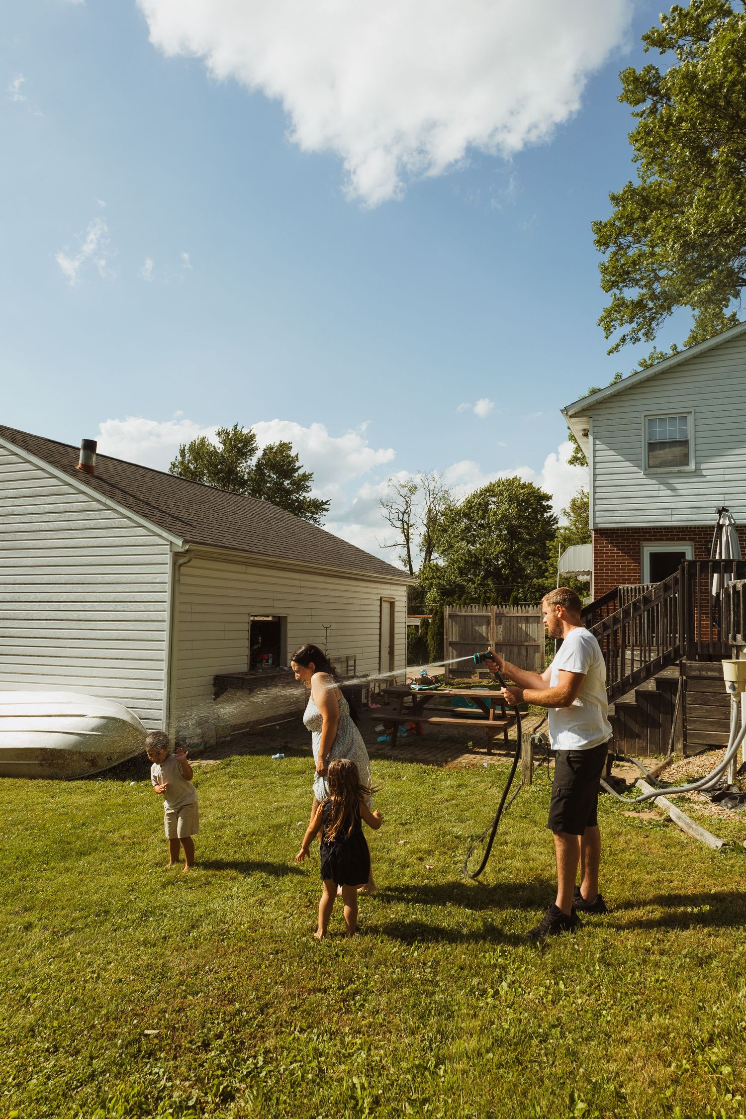 Family enjoying backyard fun with a garden hose on a sunny day near suburban homes.
