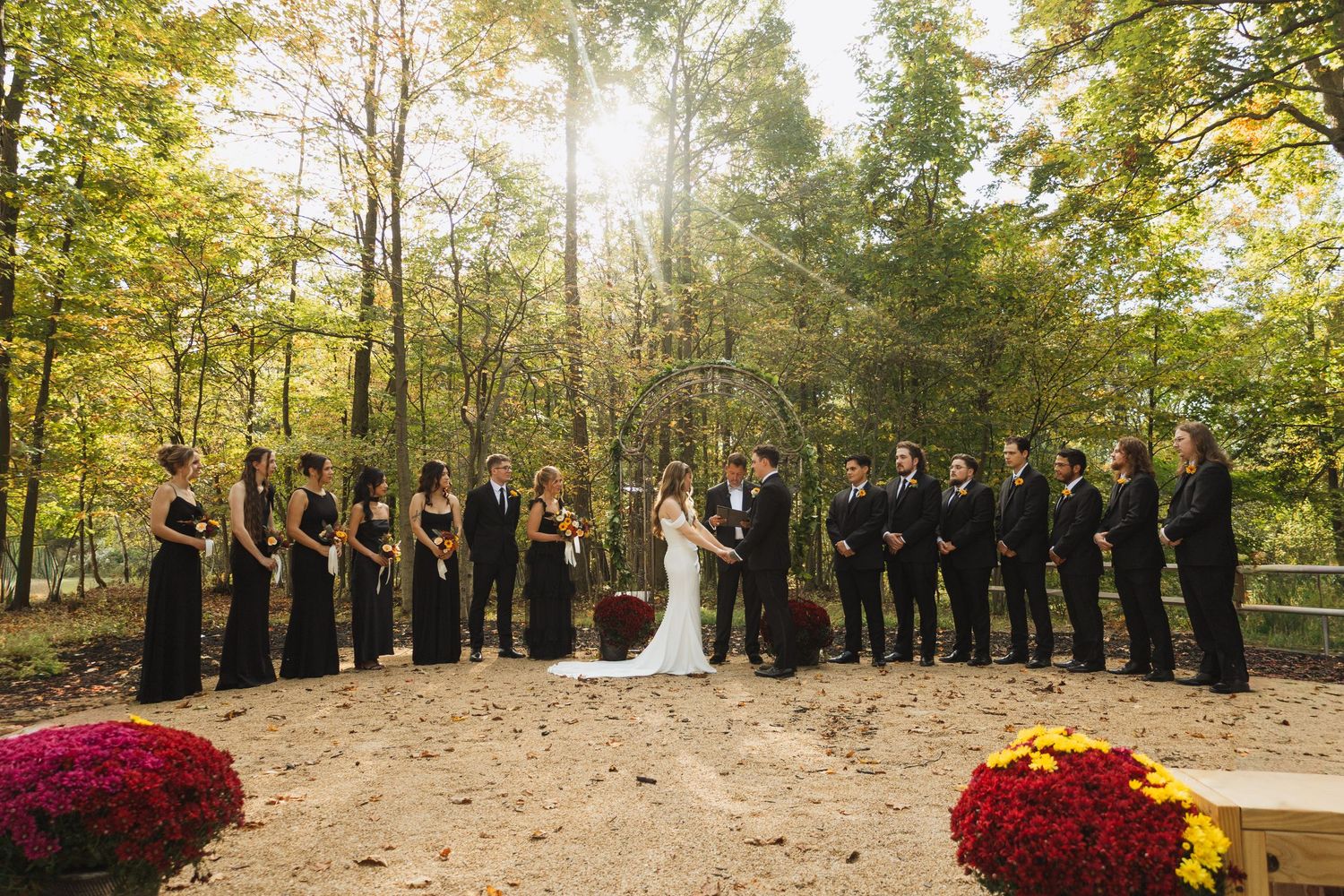 Wedding party lined up outdoors in autumn setting with vibrant red flowers and fall foliage.