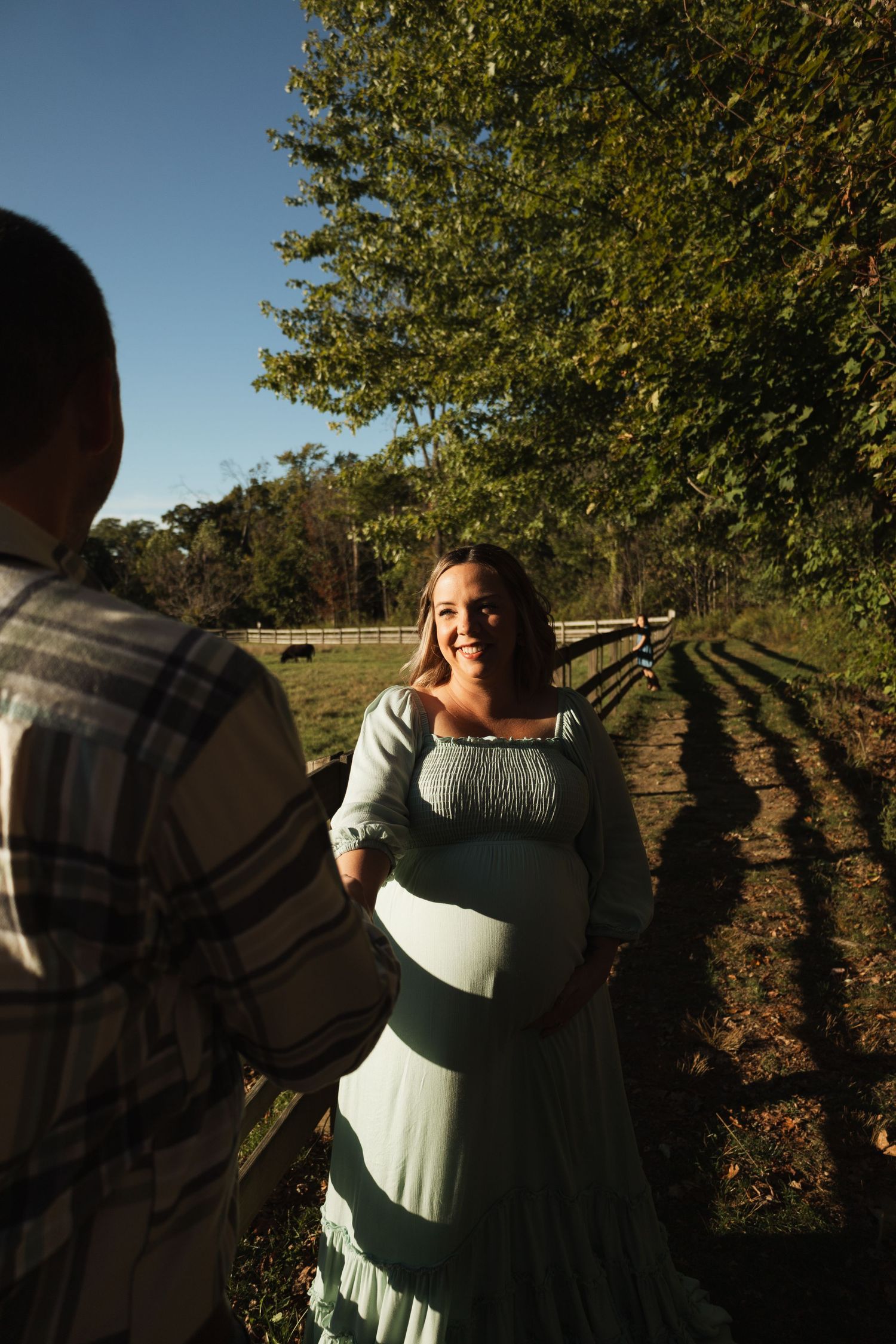 A couple dances together in a romantic outdoor setting with sunlit trees and a wooden fence in the background.