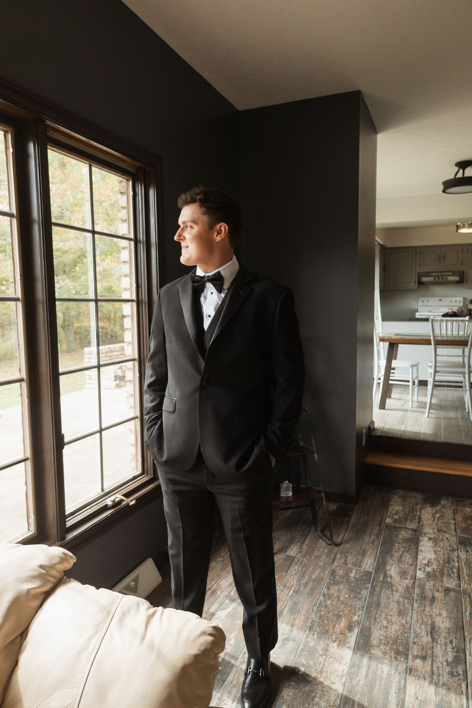 Person in formal attire standing by large windows in a modern room with wooden floors.