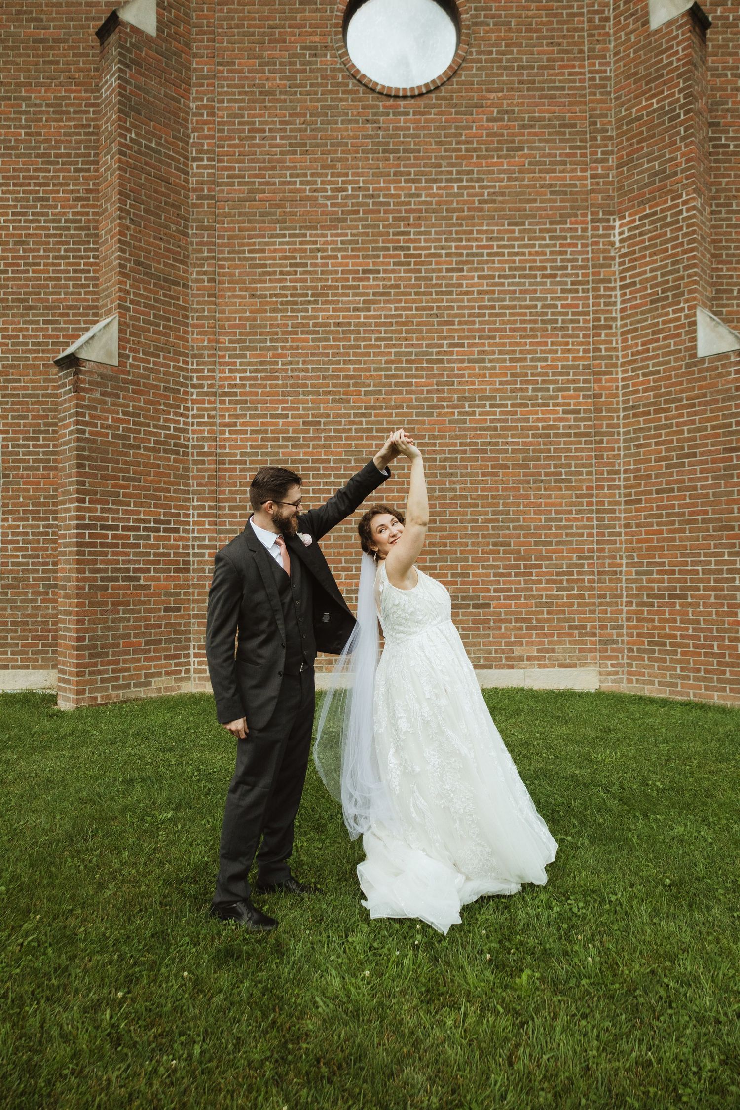 A couple dances on the lawn in front of a brick church wall during their wedding celebration.