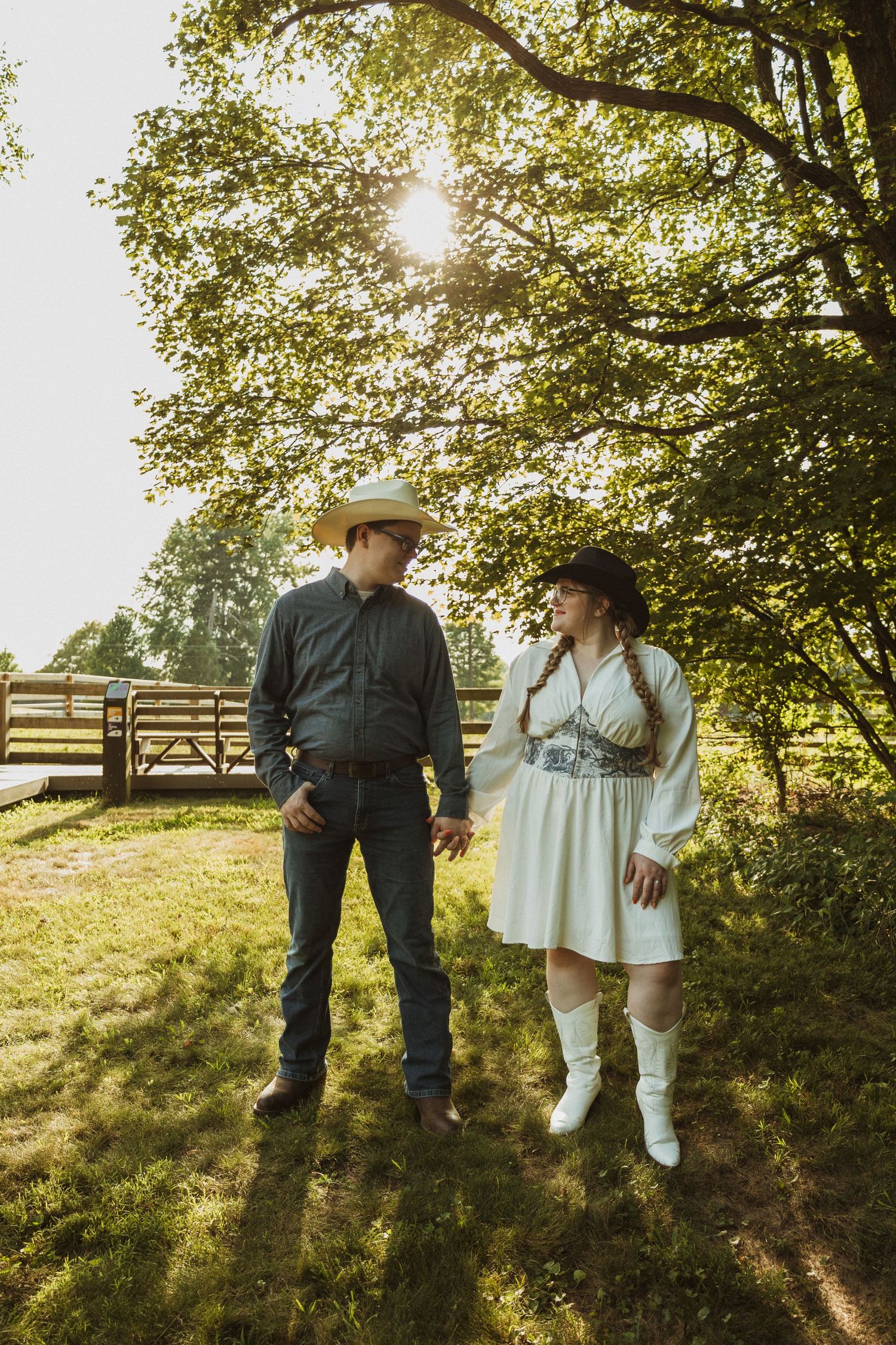 Couple in rustic attire enjoying a romantic moment on a sunlit grassy hillside with trees in the background.