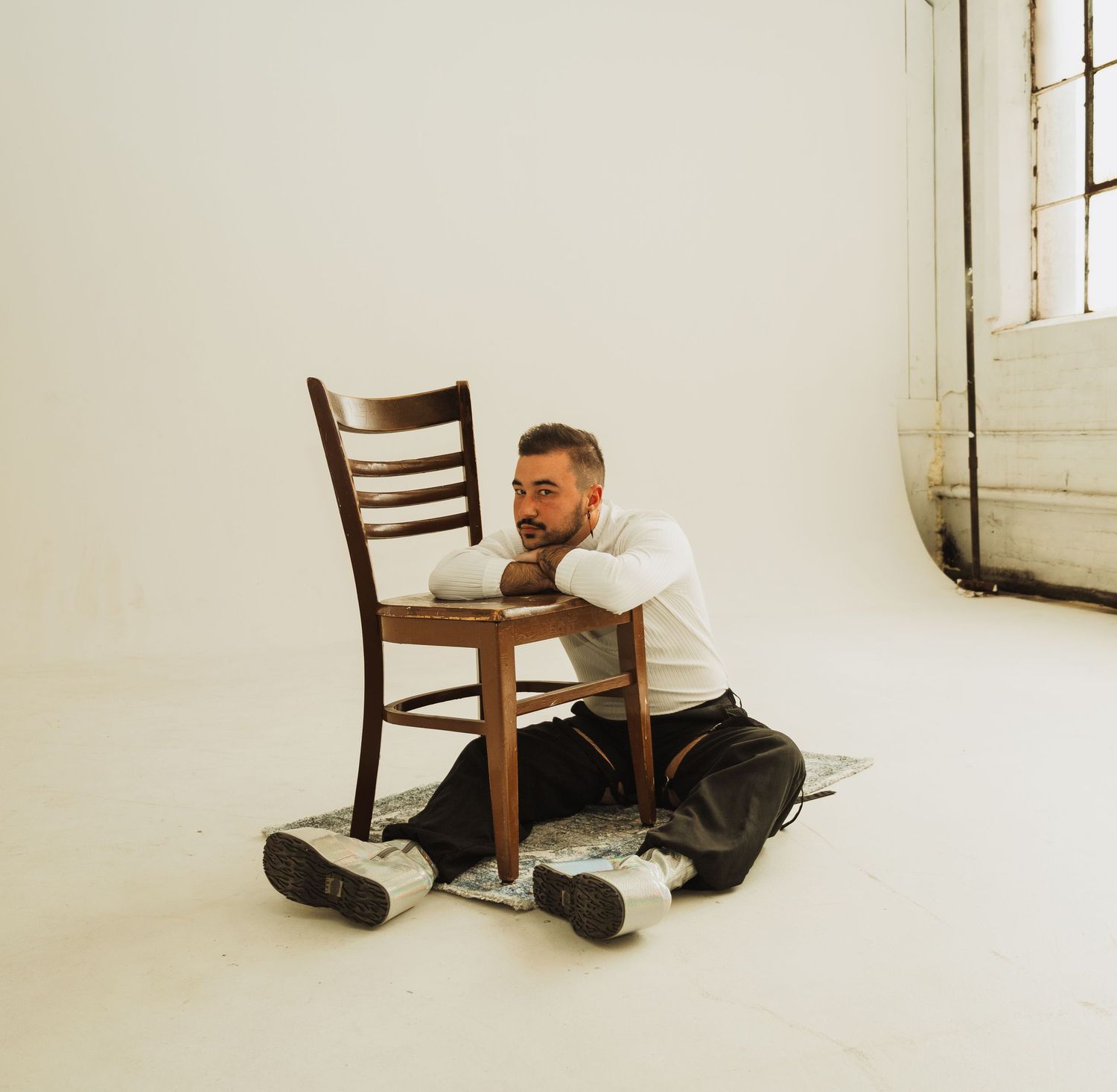 Person in white shirt and dark pants sitting on floor next to wooden chair in minimal white studio setting.