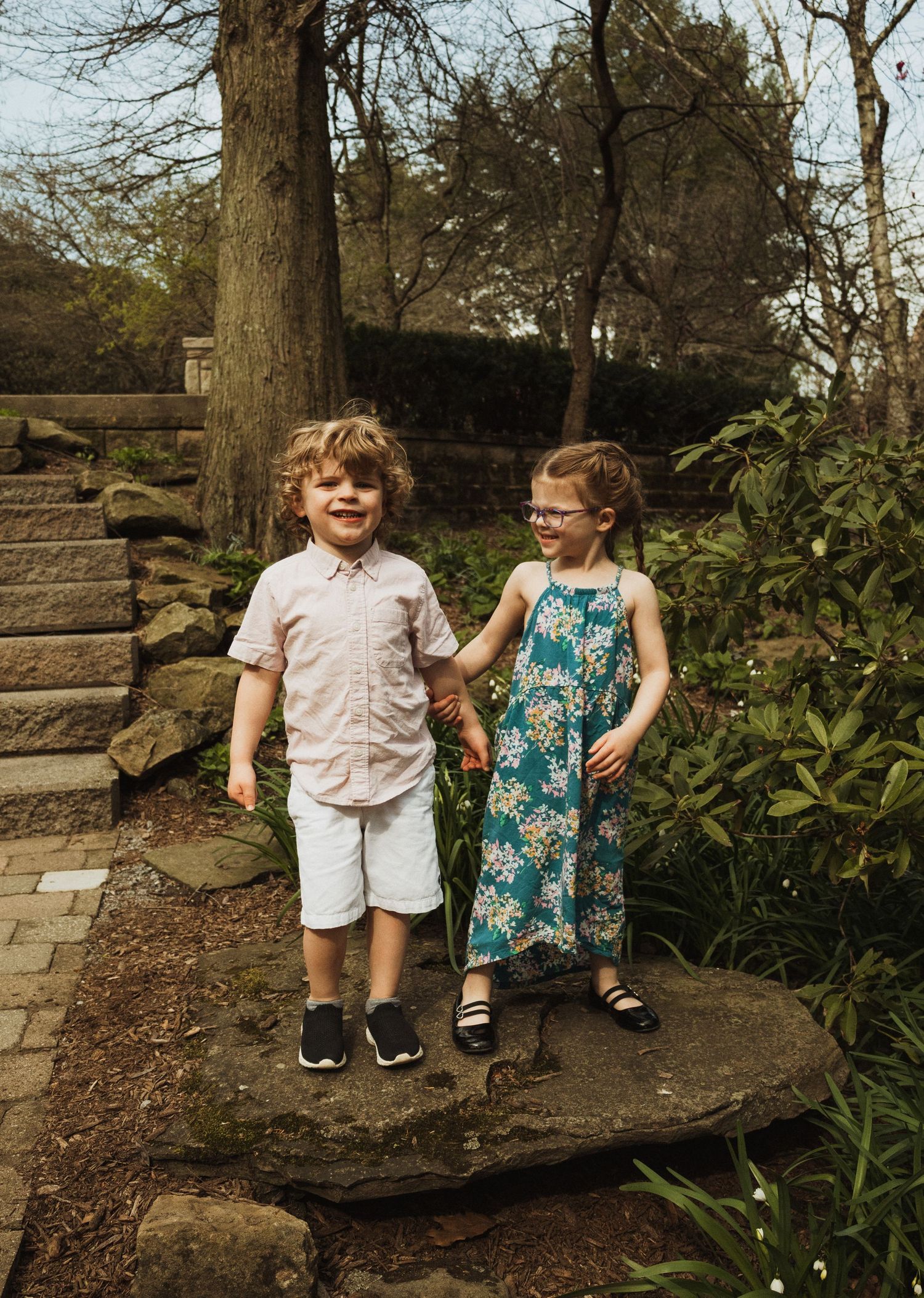 Two children stand together on stone steps in a garden path surrounded by trees and greenery.