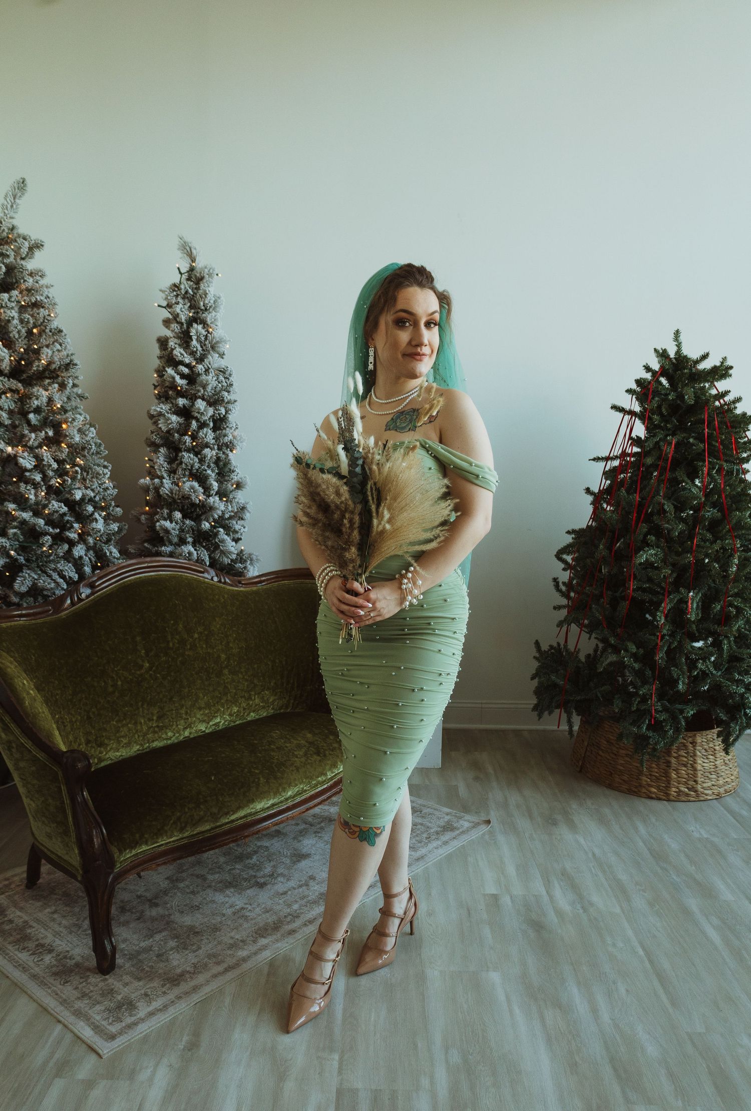 Bride in sage green lace dress with teal veil holding dried bouquet near Christmas trees.