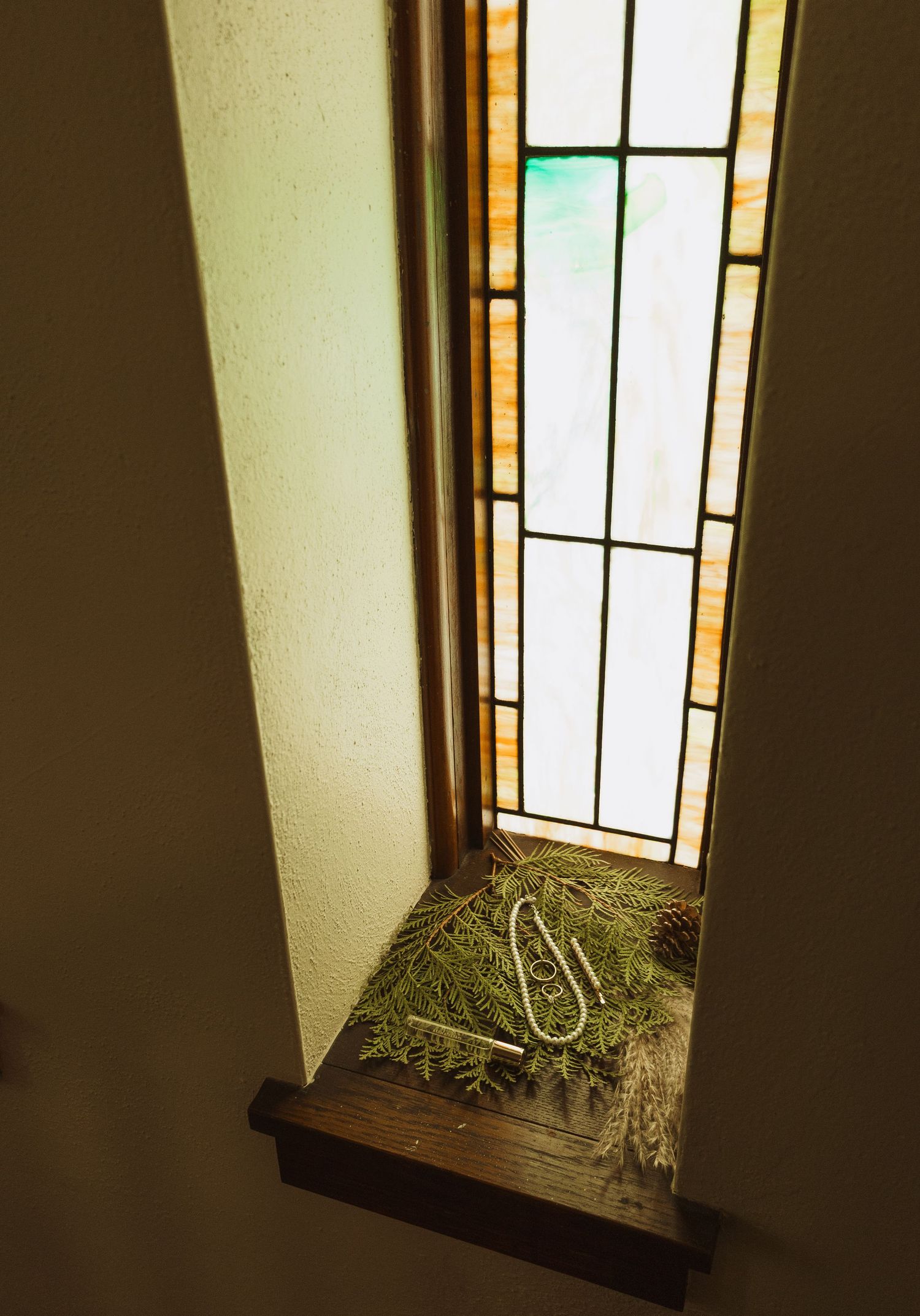 Pine branches and pearl necklace arranged on wooden window sill beside tall stained glass window.