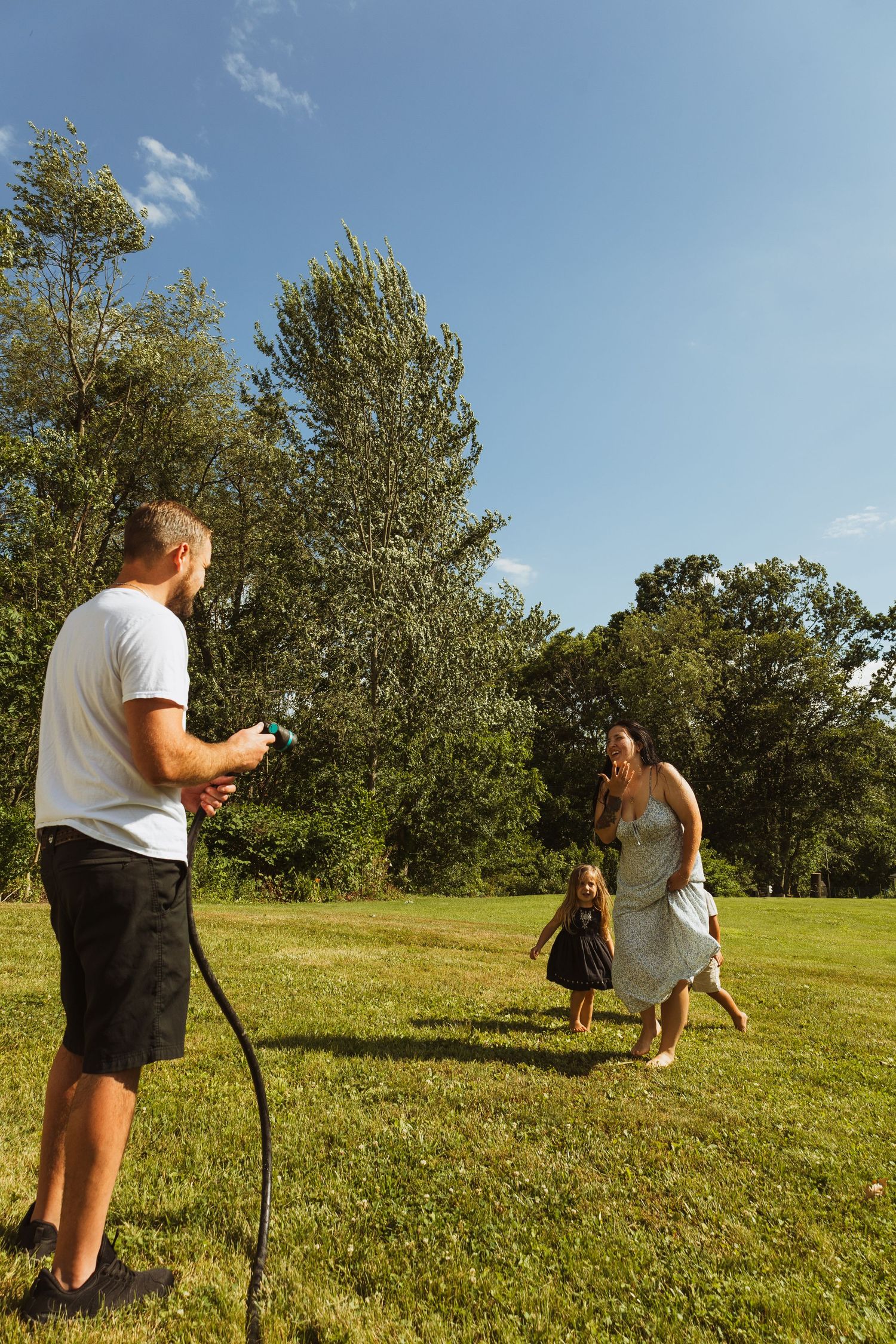 Family playing with garden hose on sunny green lawn, man spraying water at laughing woman and child outdoors.