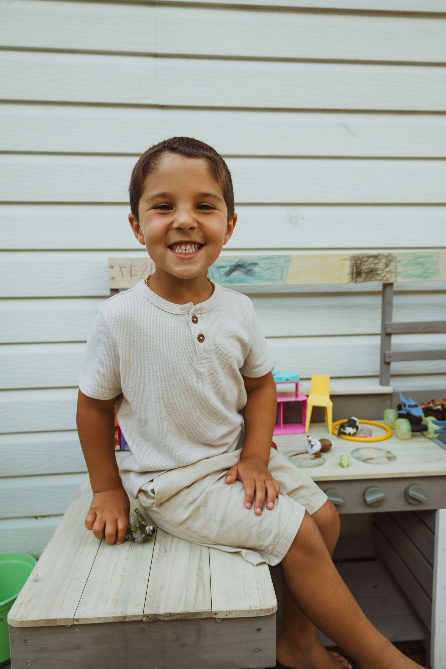 Smiling young boy sitting on a wooden box outdoors near a play table with colorful toys against a white wall.