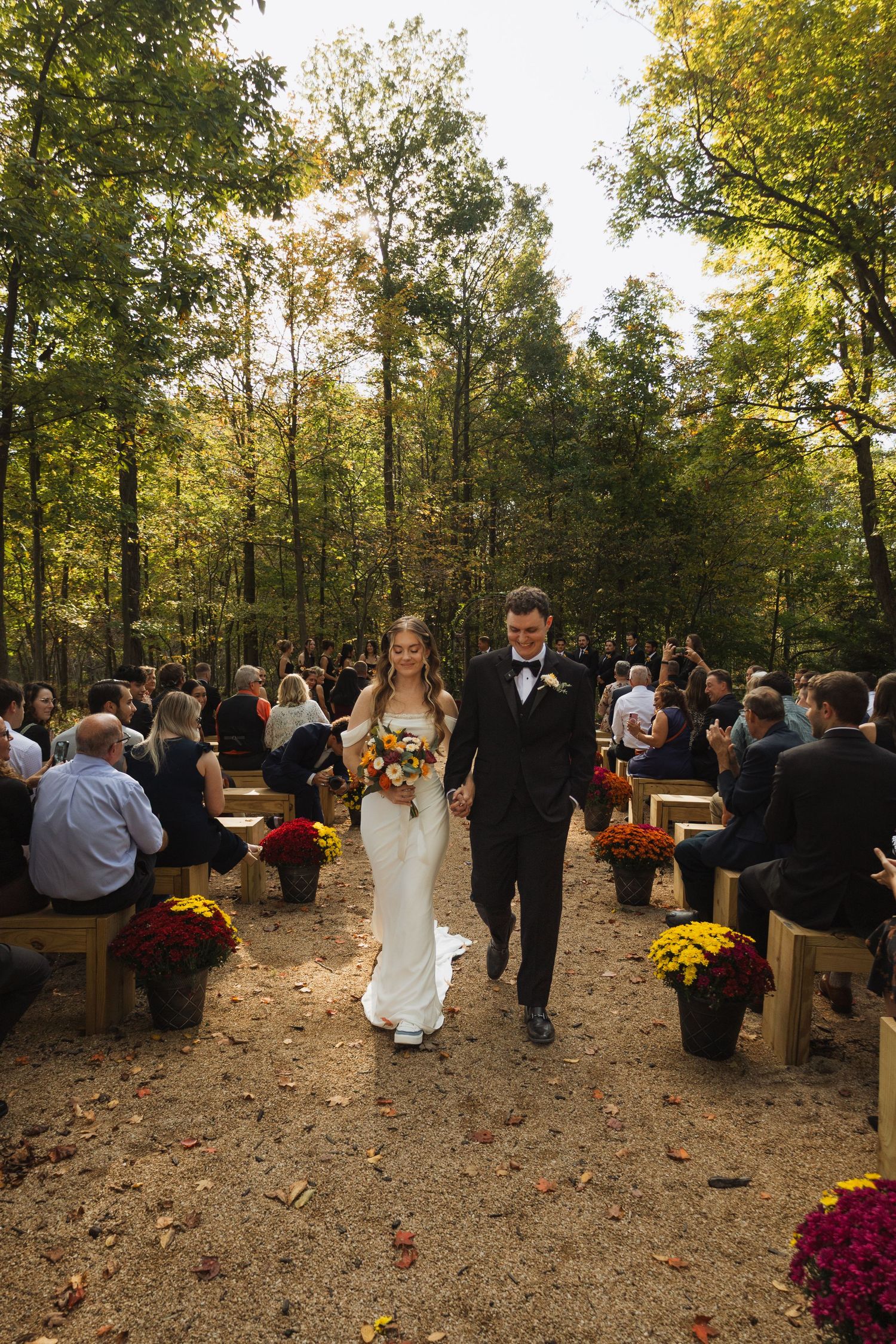 Outdoor wedding ceremony in a wooded area with wooden benches and fall mums decorating the aisle.