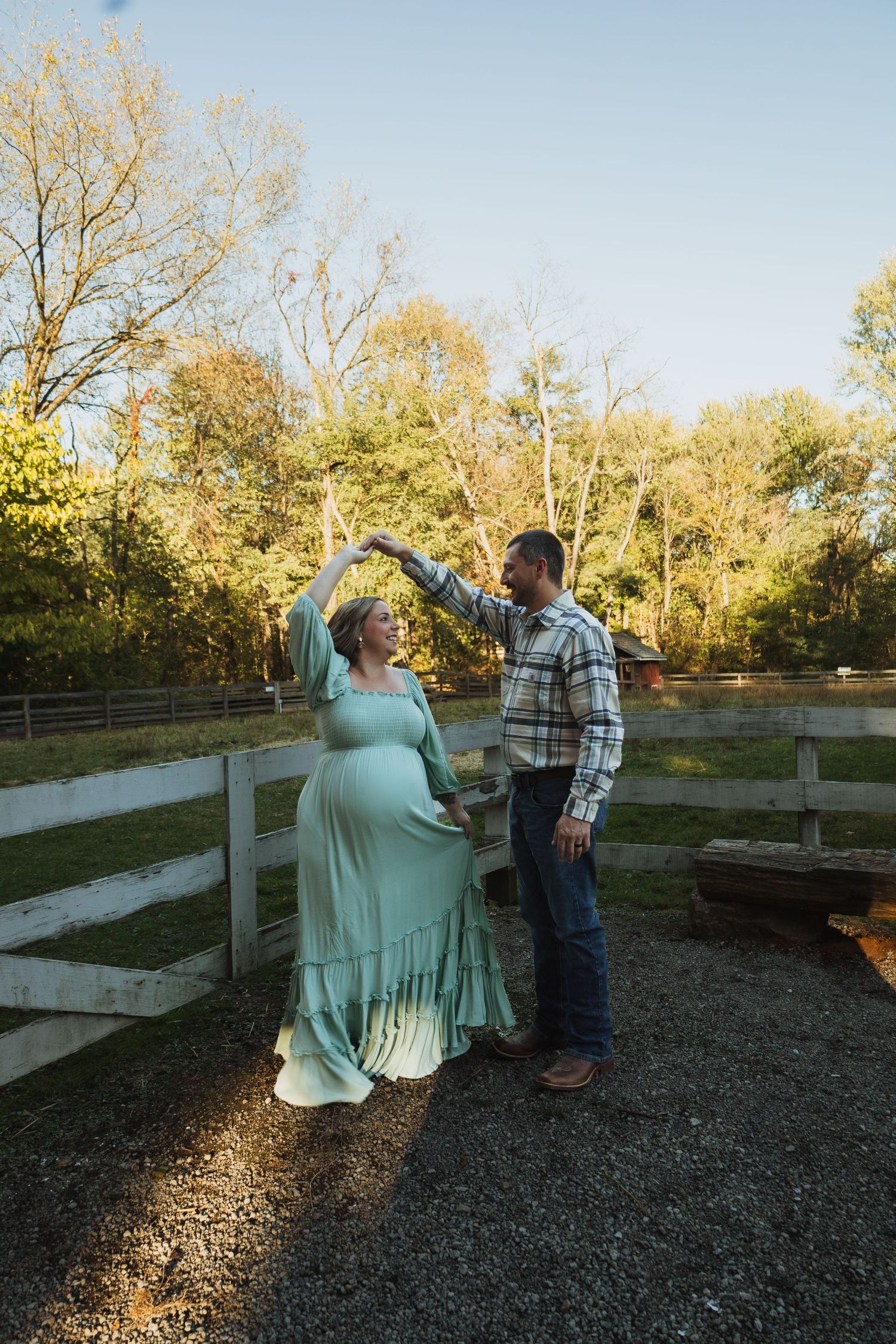 A couple stands together in a rustic outdoor setting during golden hour with wooden fencing in the background.
