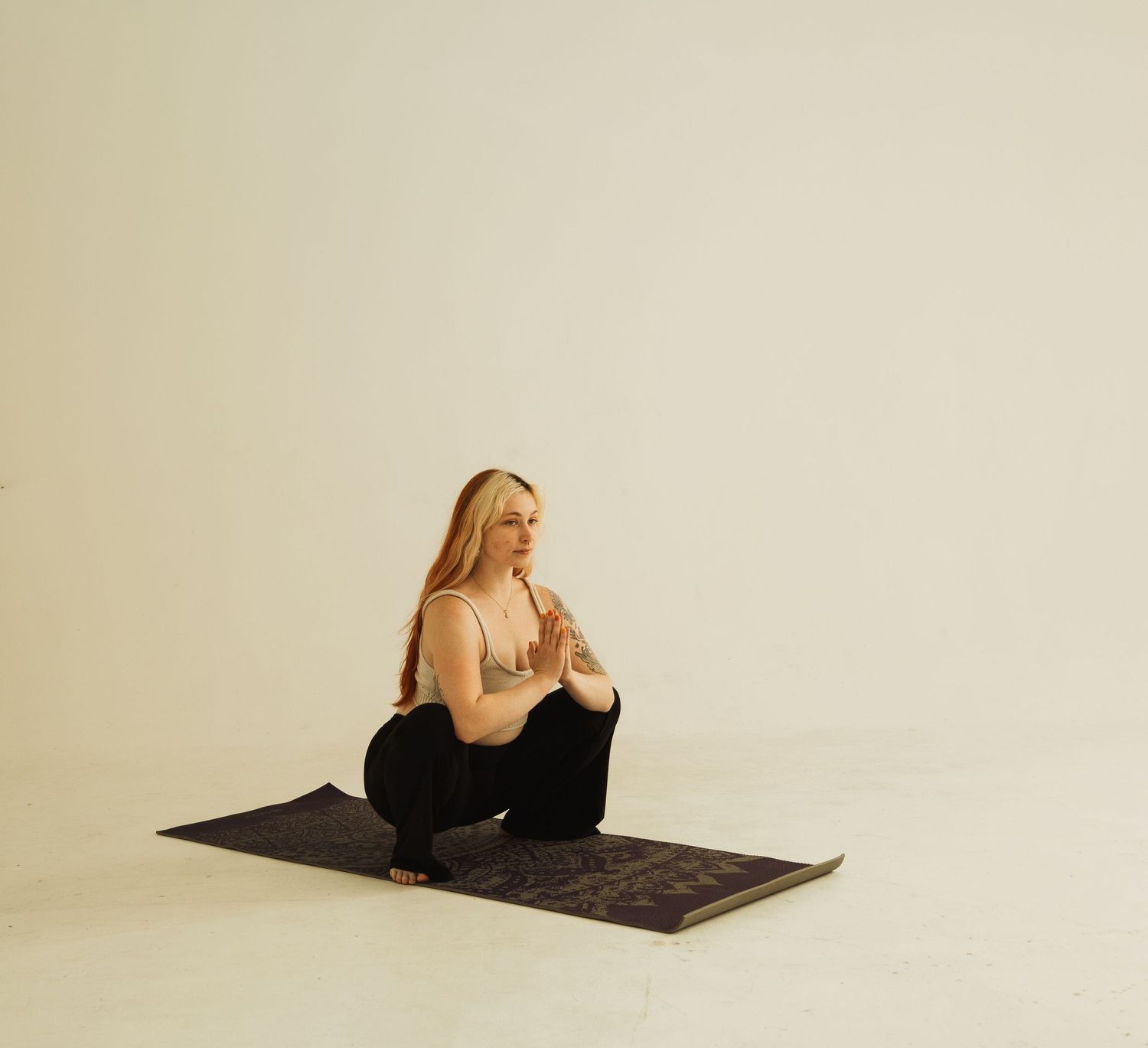 Person seated in meditation pose on black yoga mat against neutral background wearing light top and dark pants.