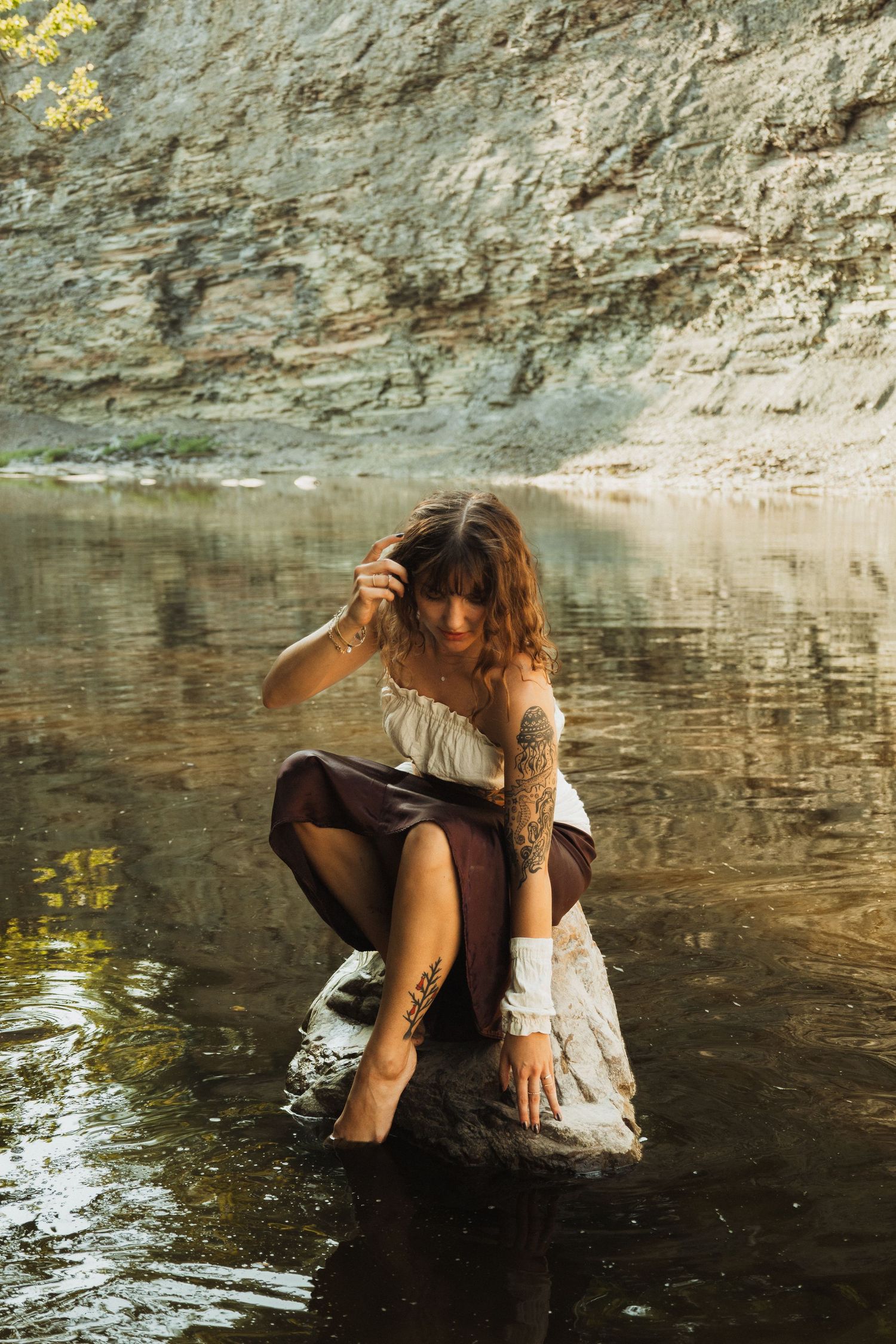 Person in casual attire lounging by a calm reflective stream against a rocky cliff face.
