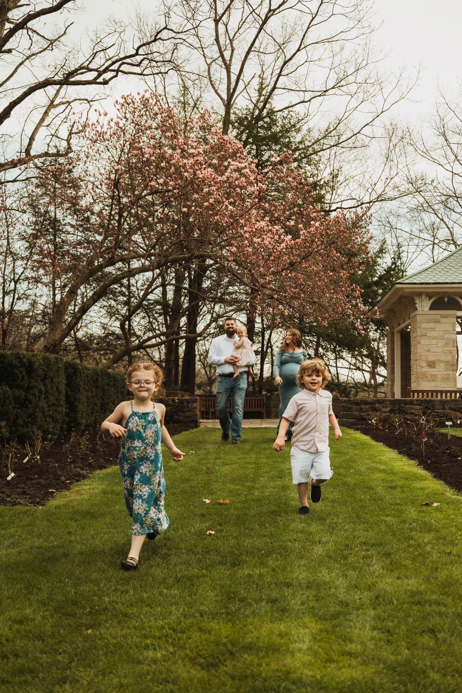 Children running and playing on a green lawn with blooming cherry trees and a stone gazebo in the background.