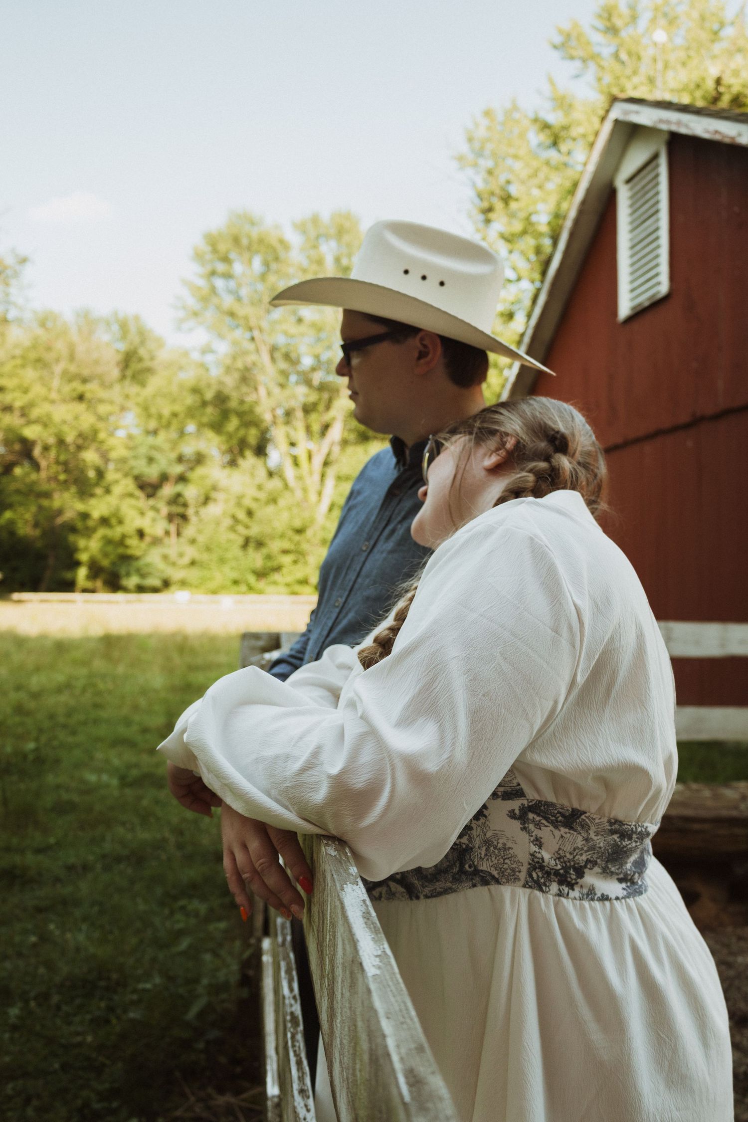 Couple in vintage-inspired clothing sitting together near a red barn in warm afternoon light.