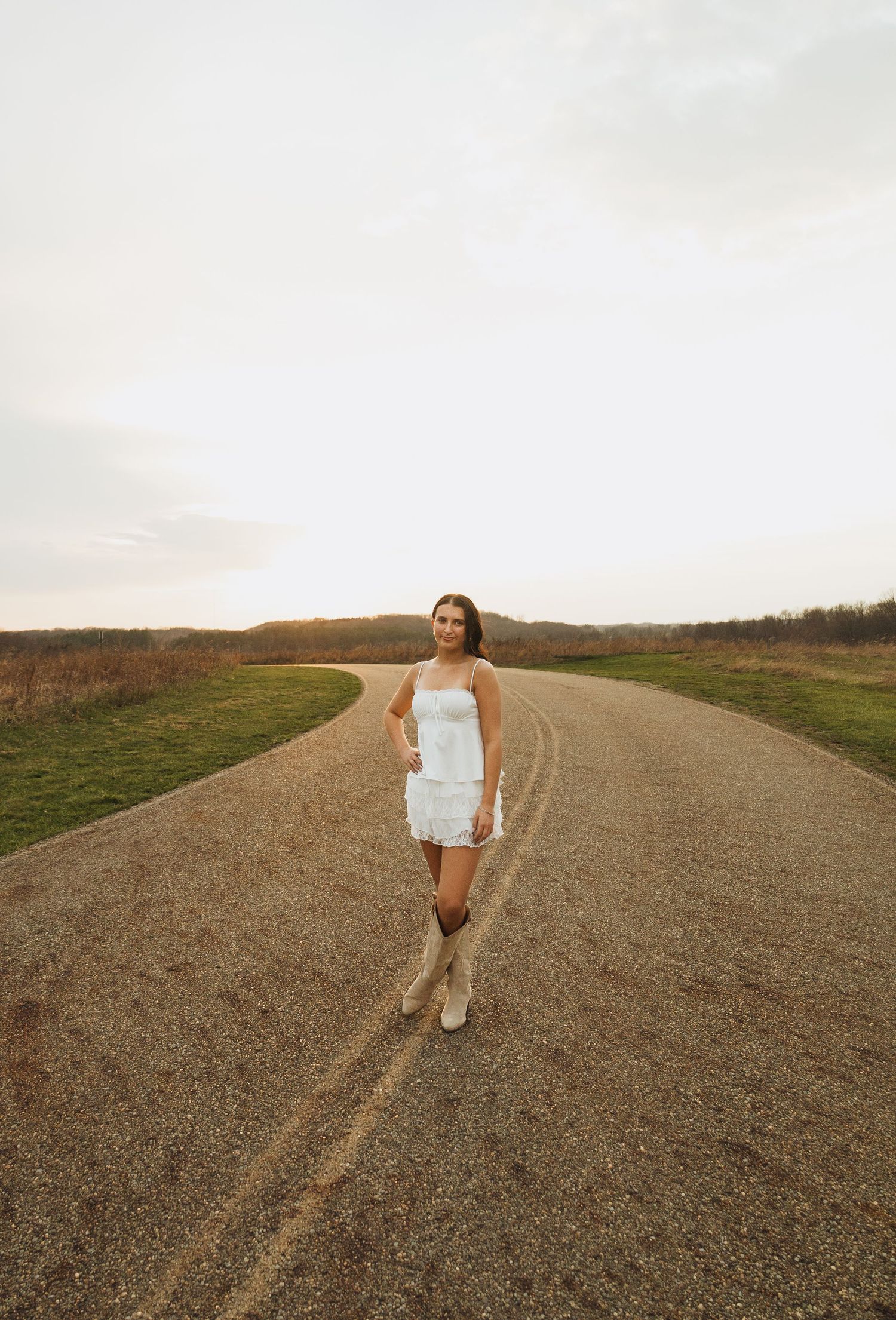 Woman in white dress and cowboy boots standing on a rural dirt road surrounded by open fields.